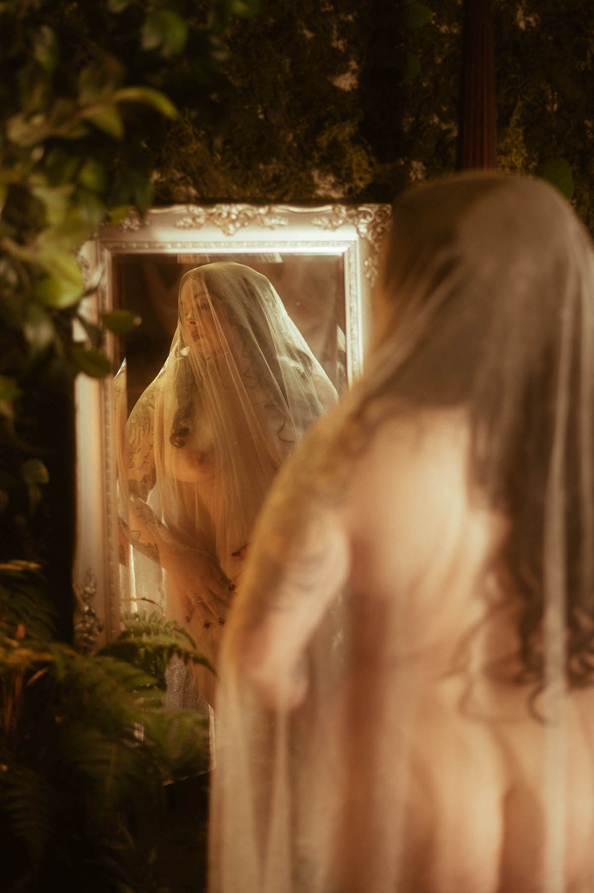 A nude figure draped in a sheer veil gazes into an ornate mirror at Royal Lune Studio, surrounded by ferns and a textured moss wall. This moody boudoir photography scene blends fantasy and fine art, evoking dreamy bridal and goddess-inspired storytelling.