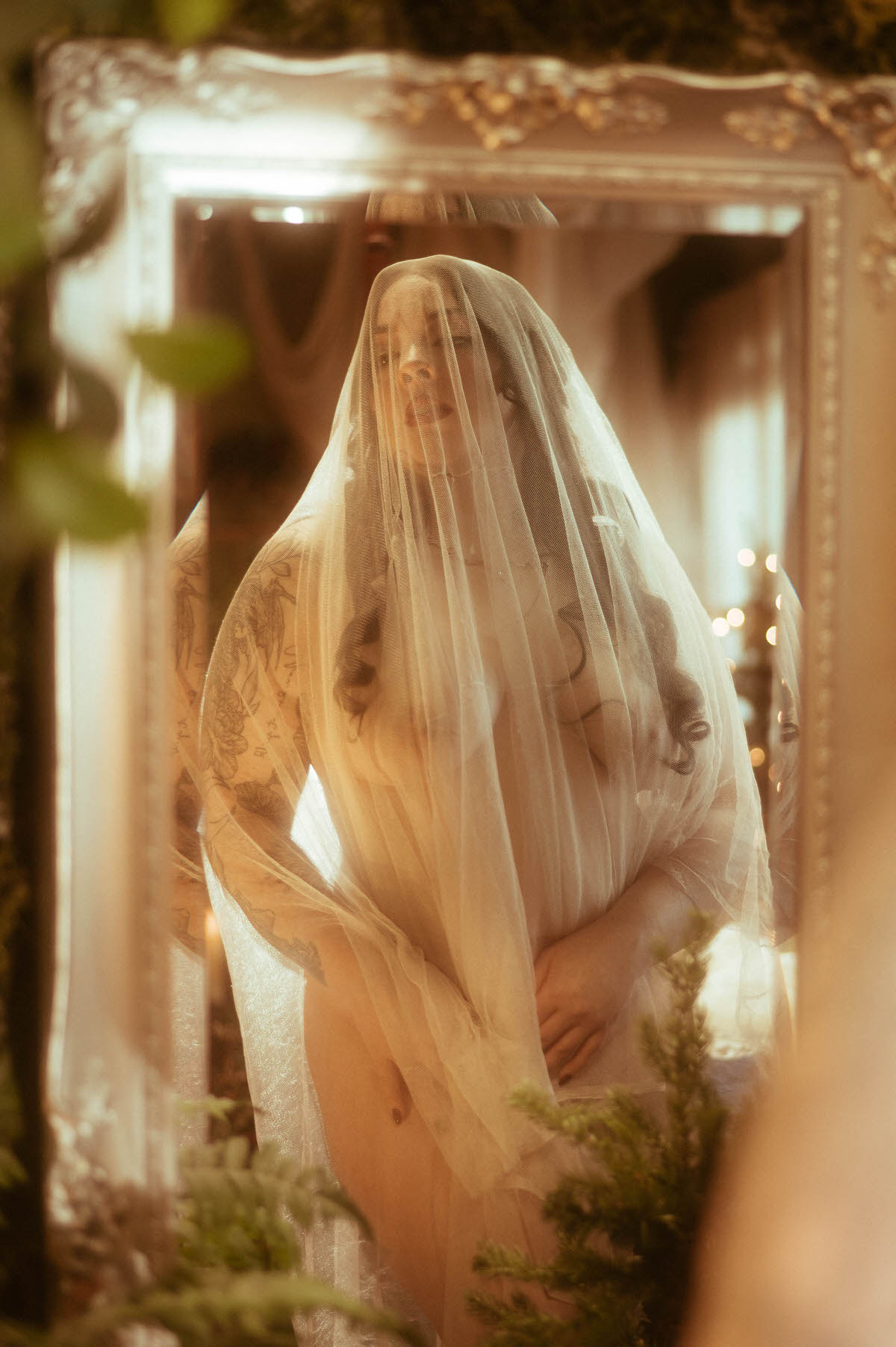A soft, romantic boudoir portrait of a nude muse draped in a sheer embroidered veil, reflected in a vintage mirror at Royal Lune Studio. Surrounded by greenery and golden light, evoking ethereal bridal and fine art fantasy themes in a moody boudoir photography style.