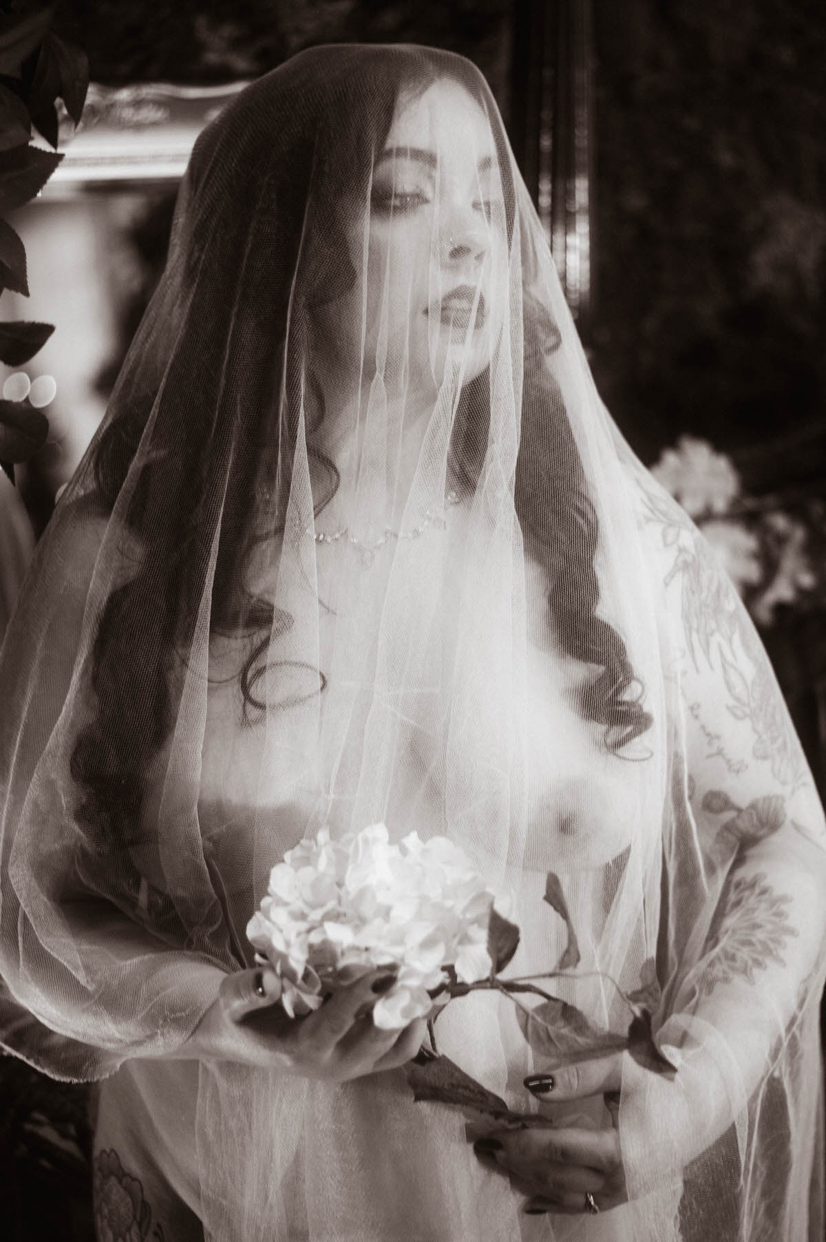 A black-and-white boudoir portrait of a veiled woman holding a single hydrangea stem, with soft curls cascading over her shoulders and lace veiling draped across her bare form. Shot at Royal Lune Studio, this image captures the poetic elegance of moody boudoir photography with a timeless, vintage atmosphere.