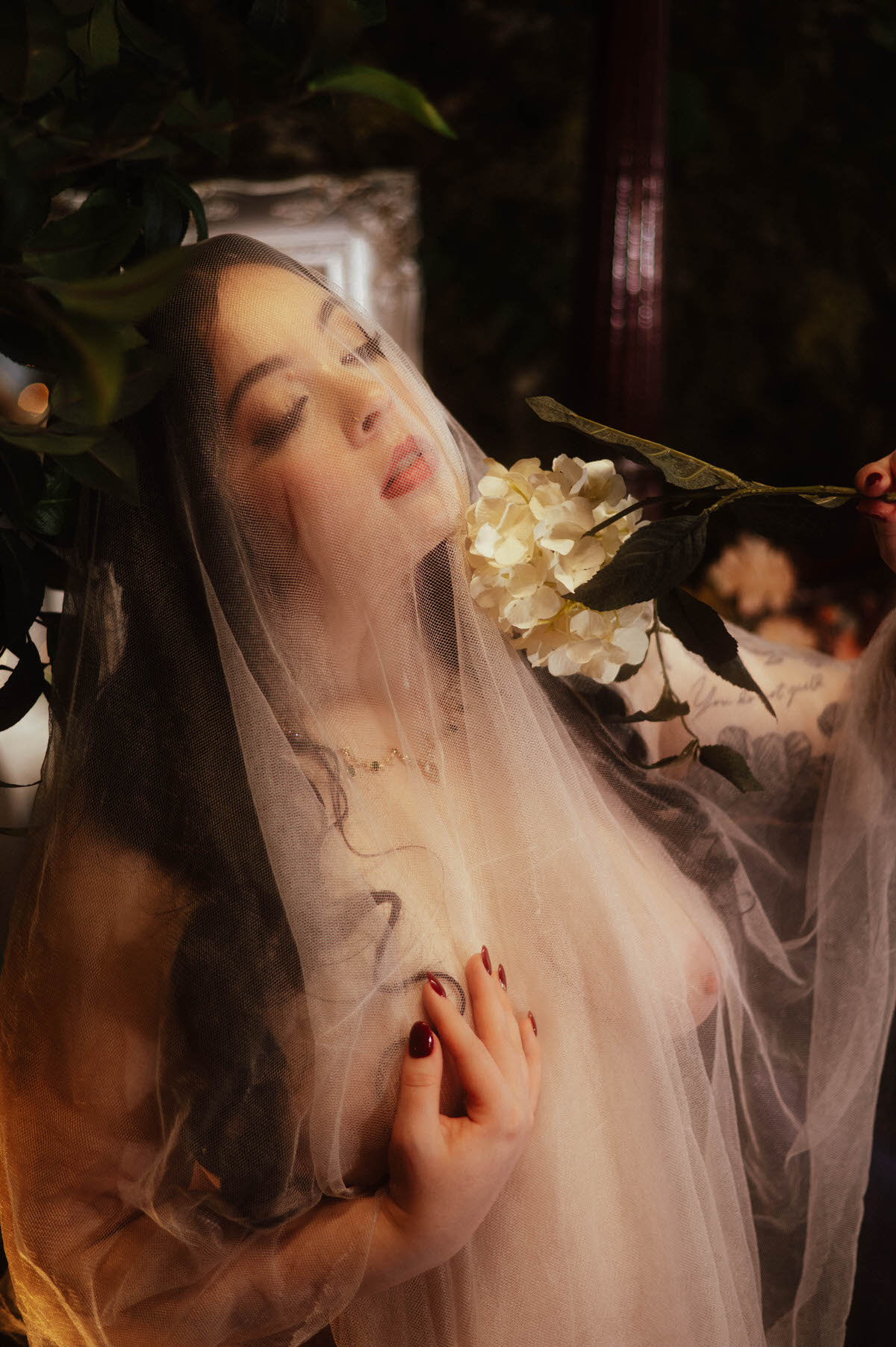 A romantic boudoir portrait featuring a veiled woman holding a white hydrangea near her face at Royal Lune Studio. Her eyes are softly closed, lips parted, and deep red nails press gently into sheer tulle, evoking moody boudoir photography with a dreamy, floral aesthetic.