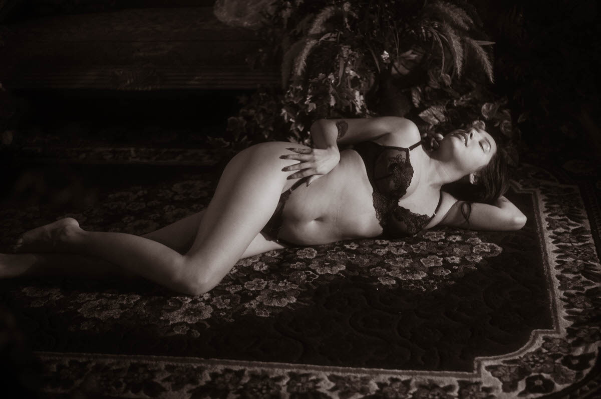 A black and white boudoir portrait of a client in lace lingerie reclining on an ornate floral rug, captured for a moody artistic nude photography session. This timeless dramatic image was created at a Dallas area studio. Royal Lune Photo.