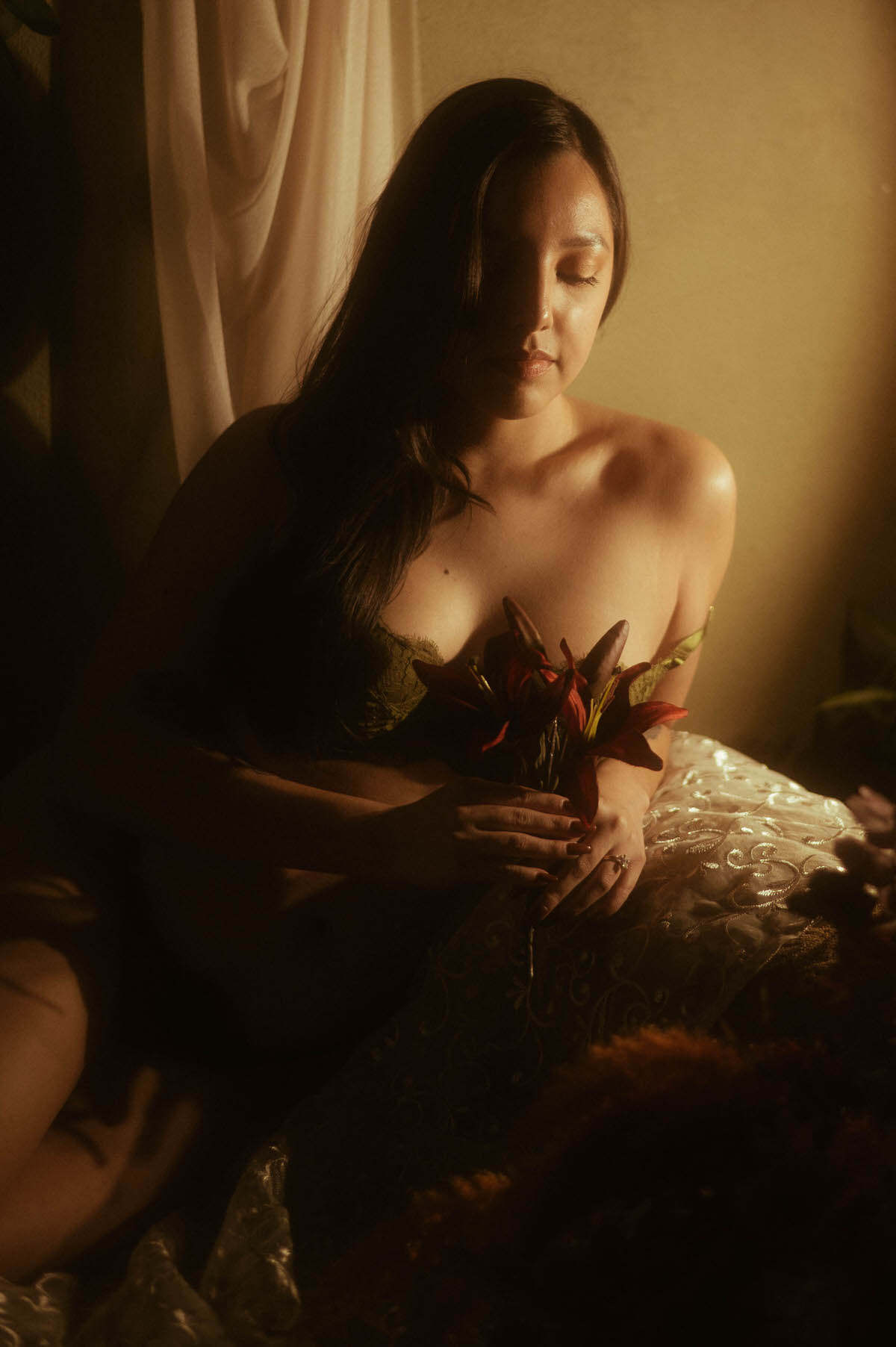 A client poses in dark green lace lingerie holding deep red lilies, captured in moody warm lighting for an intimate boudoir session. This dramatic boudoir inspiration image was created at a Dallas area studio. Royal Lune Photo.