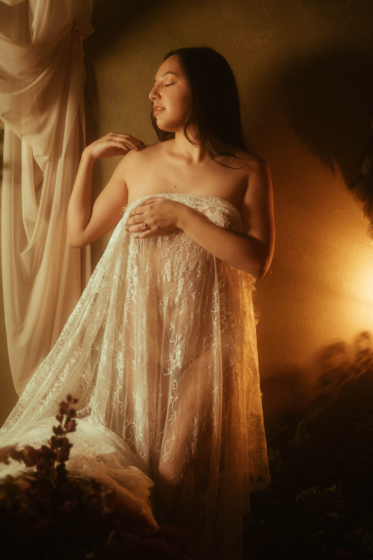 Woman draped in delicate lace fabric beside a softly lit window during a moody boudoir photos session at Royal Lune Studio. Warm golden light and dramatic shadows create fine art boudoir photography inspiration by a Dallas boudoir photographer.