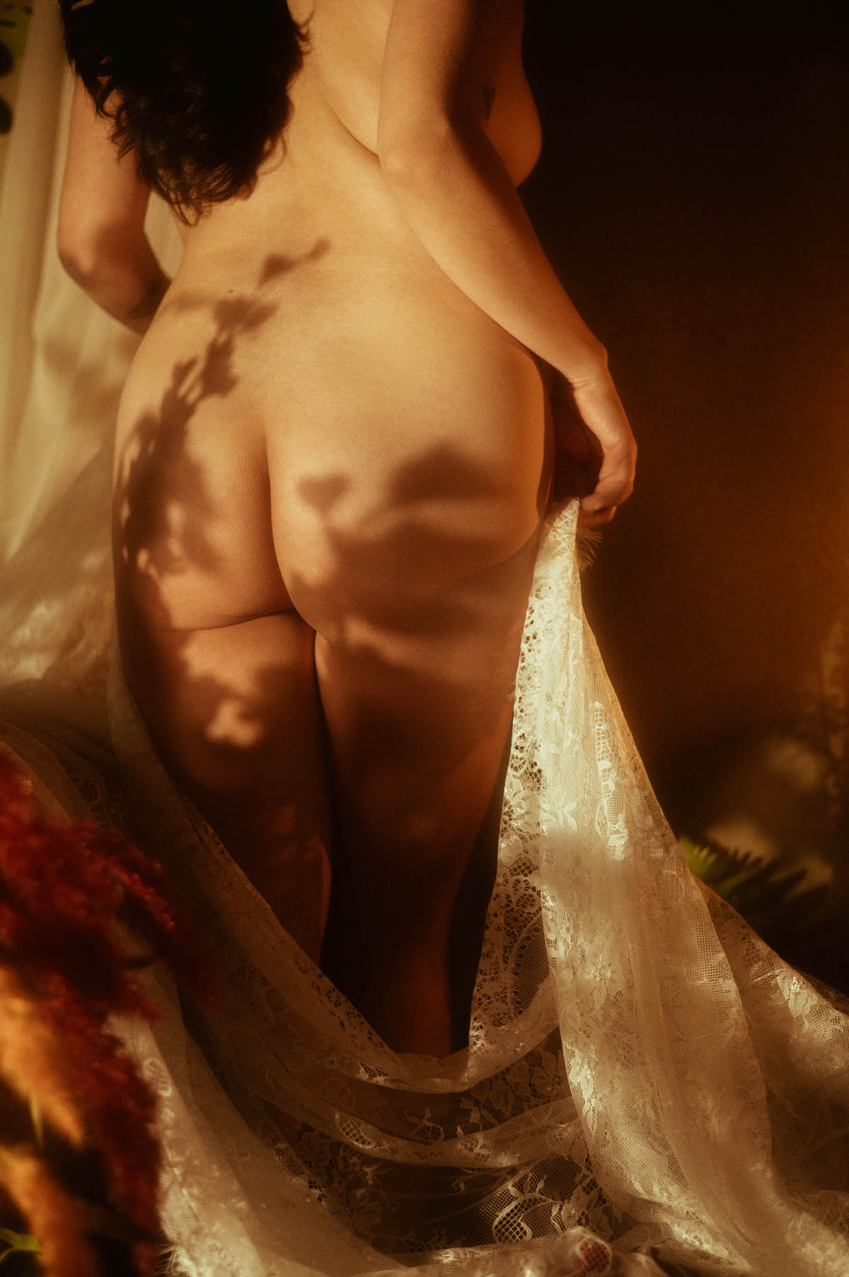 A moody boudoir photo showcasing soft dappled shadows across the body with sheer lace draped at the hips, captured in warm low lighting for artistic nude photography. This dark and romantic image reflects the work of a Dallas boudoir photographer. Royal Lune Photo.