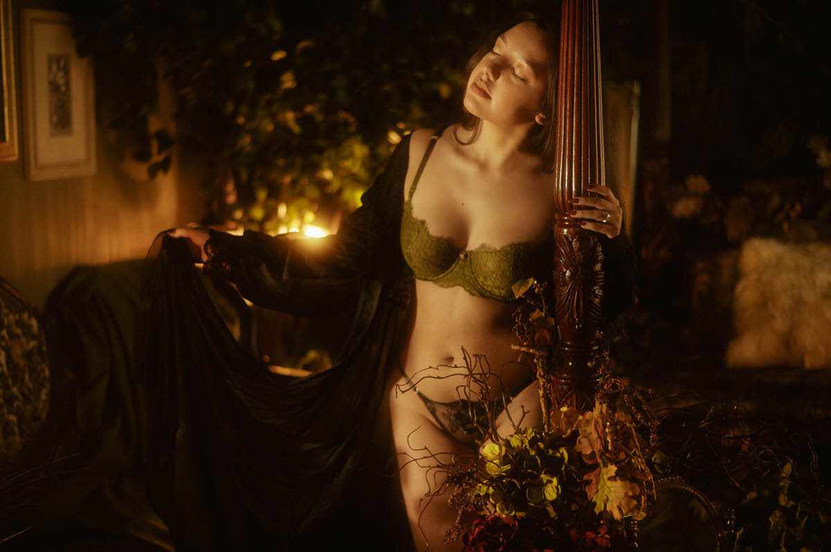 A client in dark green lace lingerie leans against a carved wooden bedpost surrounded by dried florals and warm candlelight, captured for a dramatic boudoir session. This moody artistic nude photography image reflects the work of a Dallas boudoir photographer. Royal Lune Photo.