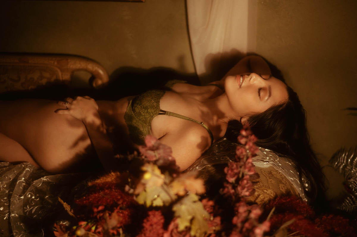 A client in dark green lace lingerie reclines among autumn foliage with soft moody lighting, captured for an artistic nude photography session. This dark romantic boudoir image was created at a Dallas area studio. Royal Lune Photo.