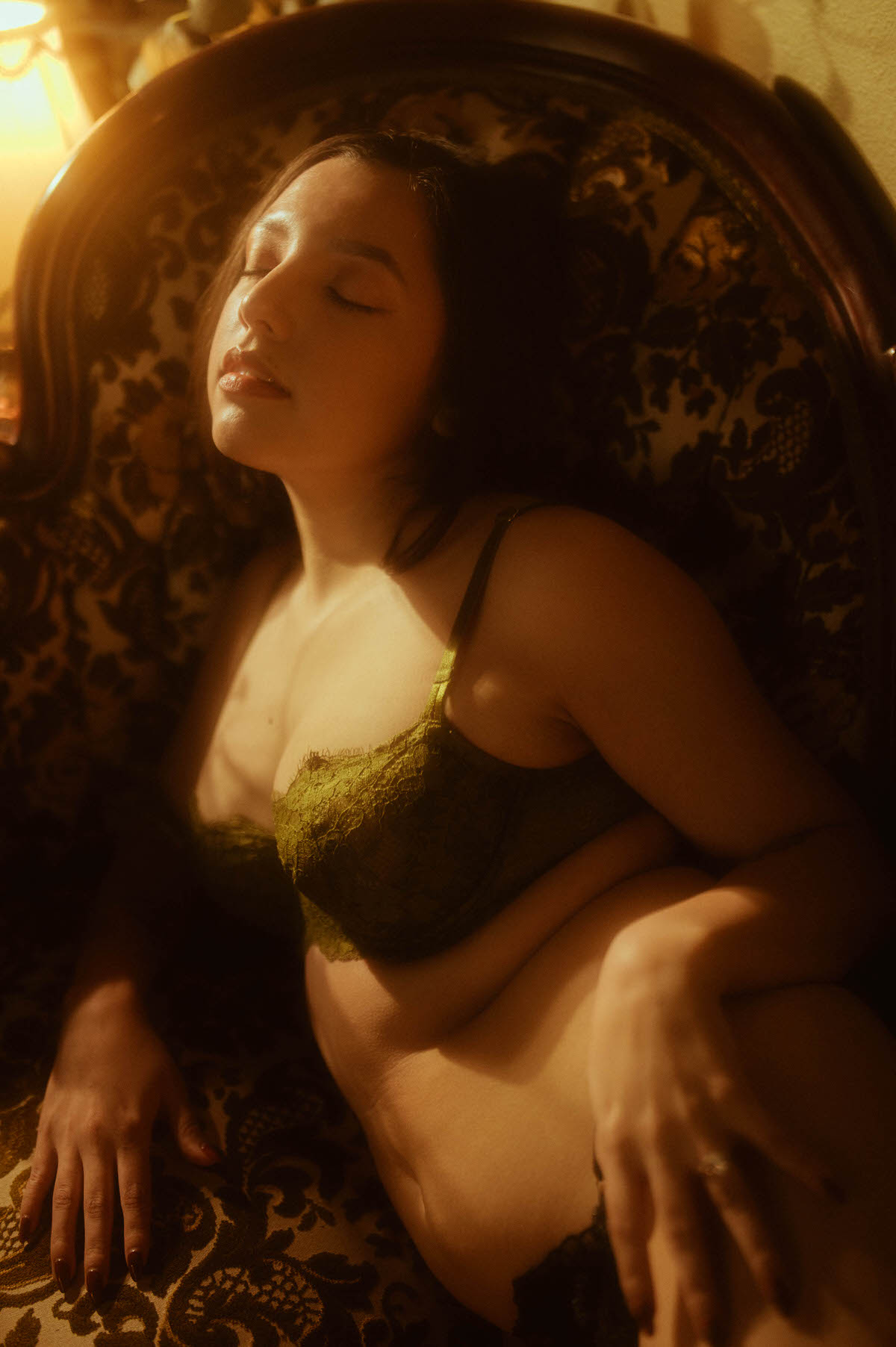 A client in dark green lace lingerie rests against an ornate vintage chair in warm dramatic lighting, captured for a moody boudoir inspiration session. This intimate fine art image was created at a Dallas area studio. Royal Lune Photo.