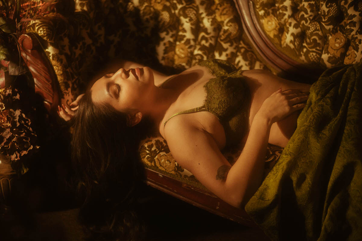 A client in dark green lace lingerie reclines against a vintage floral chaise in warm dramatic lighting, captured for a moody boudoir photo session. This intimate fine art image was created at a Dallas area studio. Royal Lune Photo.
