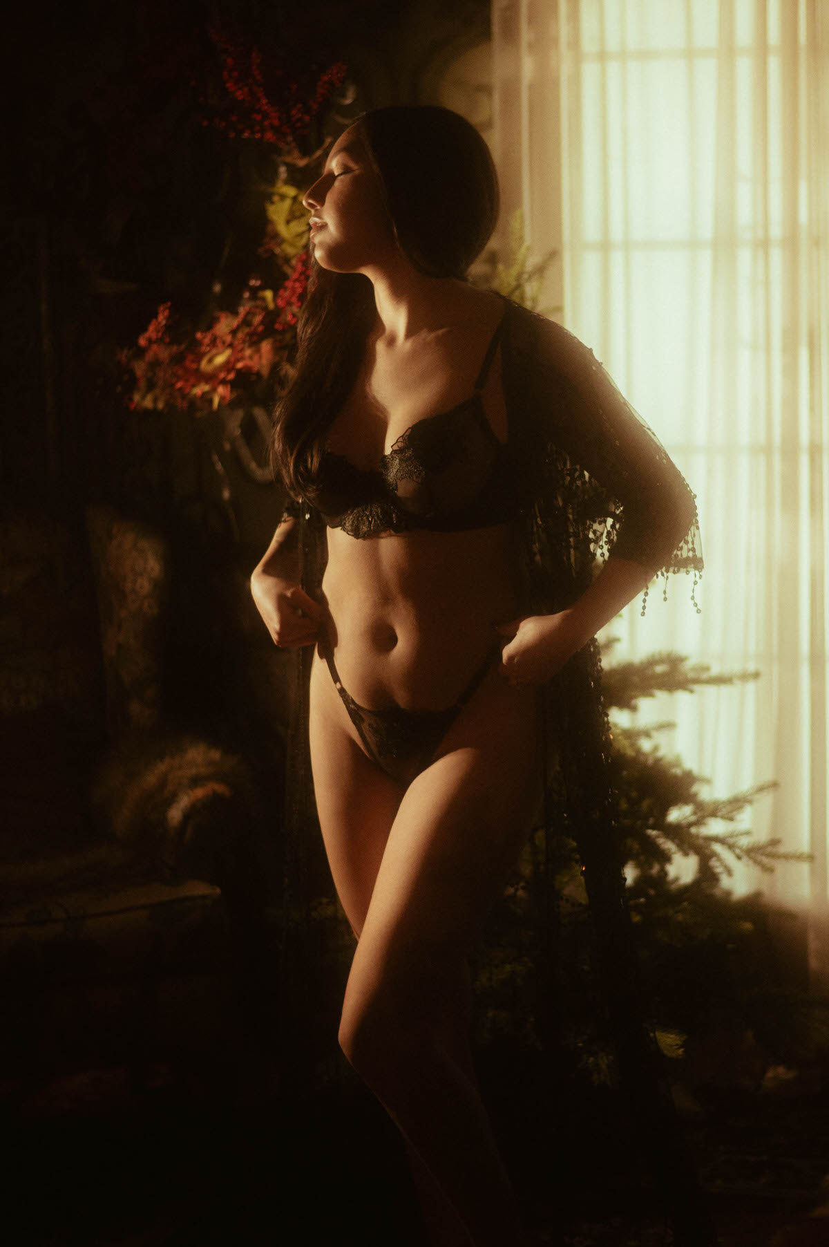 A client in black lace lingerie and a sheer fringed robe stands near a window with warm low lighting, captured for a dramatic boudoir photo session. This moody artistic nude photography image was created at a Dallas area studio. Royal Lune Photo.