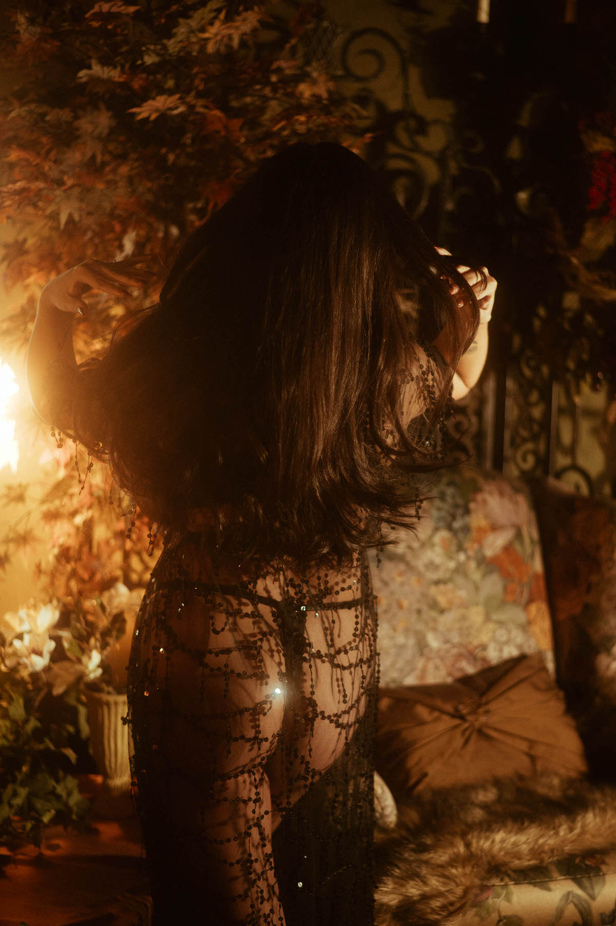 A client in a sheer black beaded robe stands among autumn foliage with warm dramatic lighting, captured from behind for an artistic nude photography session. This moody boudoir inspiration image reflects the work of a Dallas boudoir photographer. Royal Lune Photo.