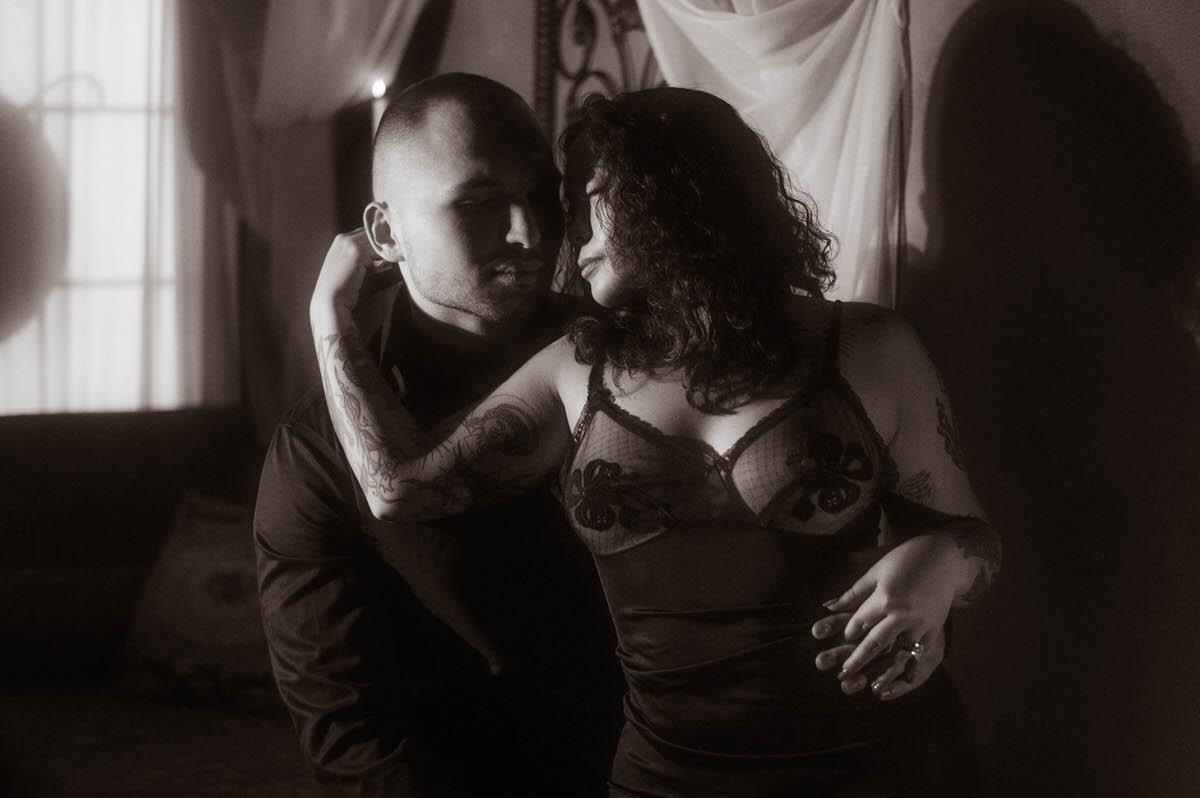 A couple shares an intense, moody gaze in this romantic couples boudoir image, with the woman in sheer black lingerie straddling her partner under candlelight shadows. Their intimate embrace captures spicy couples boudoir energy while offering bold couples boudoir ideas for emotionally charged poses.