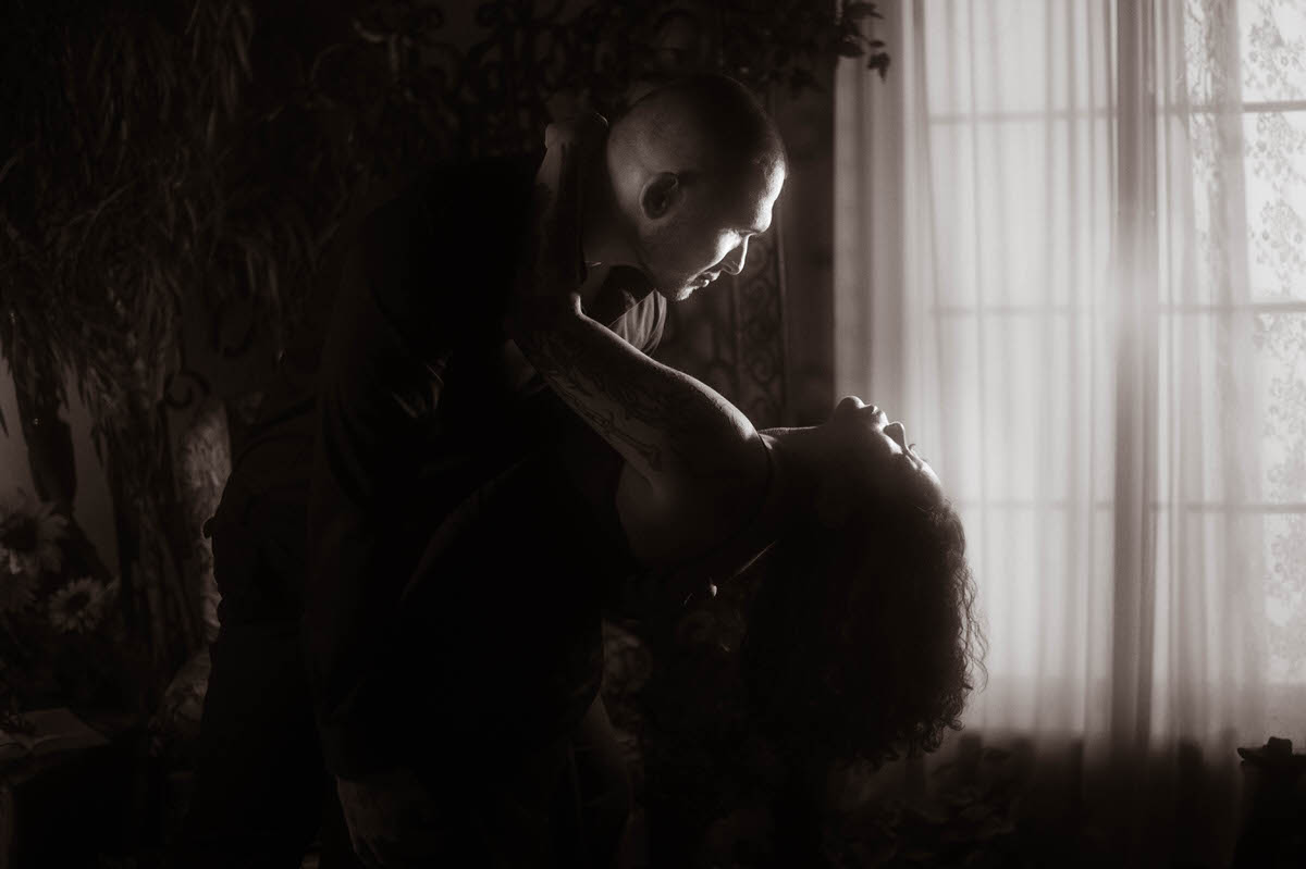 A dramatic and intimate dip pose captured in this romantic couples boudoir session, with backlighting casting soft shadows through sheer curtains. The couple’s close gaze and the moody atmosphere offer creative couples boudoir poses and a spicy couples boudoir vibe perfect for storytelling through light and shadow.