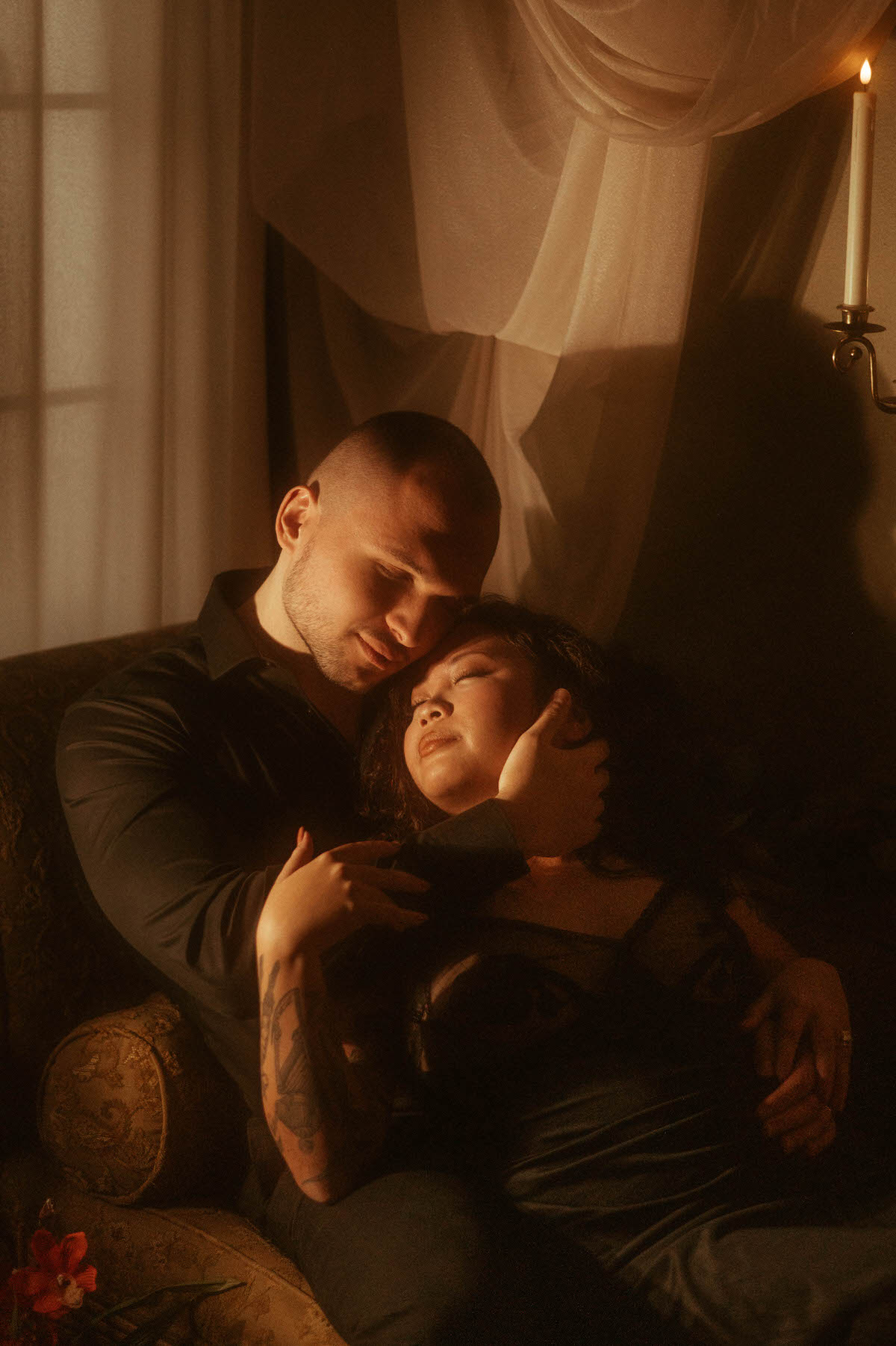 A cozy and intimate pose during a romantic couples boudoir session, with the couple curled together on a vintage armchair, bathed in soft candlelight. Her sheer black lingerie and his tender embrace highlight the emotional depth and sensuality of this moody couples boudoir moment, offering inspiration for couples boudoir poses and spicy couple photoshoots.