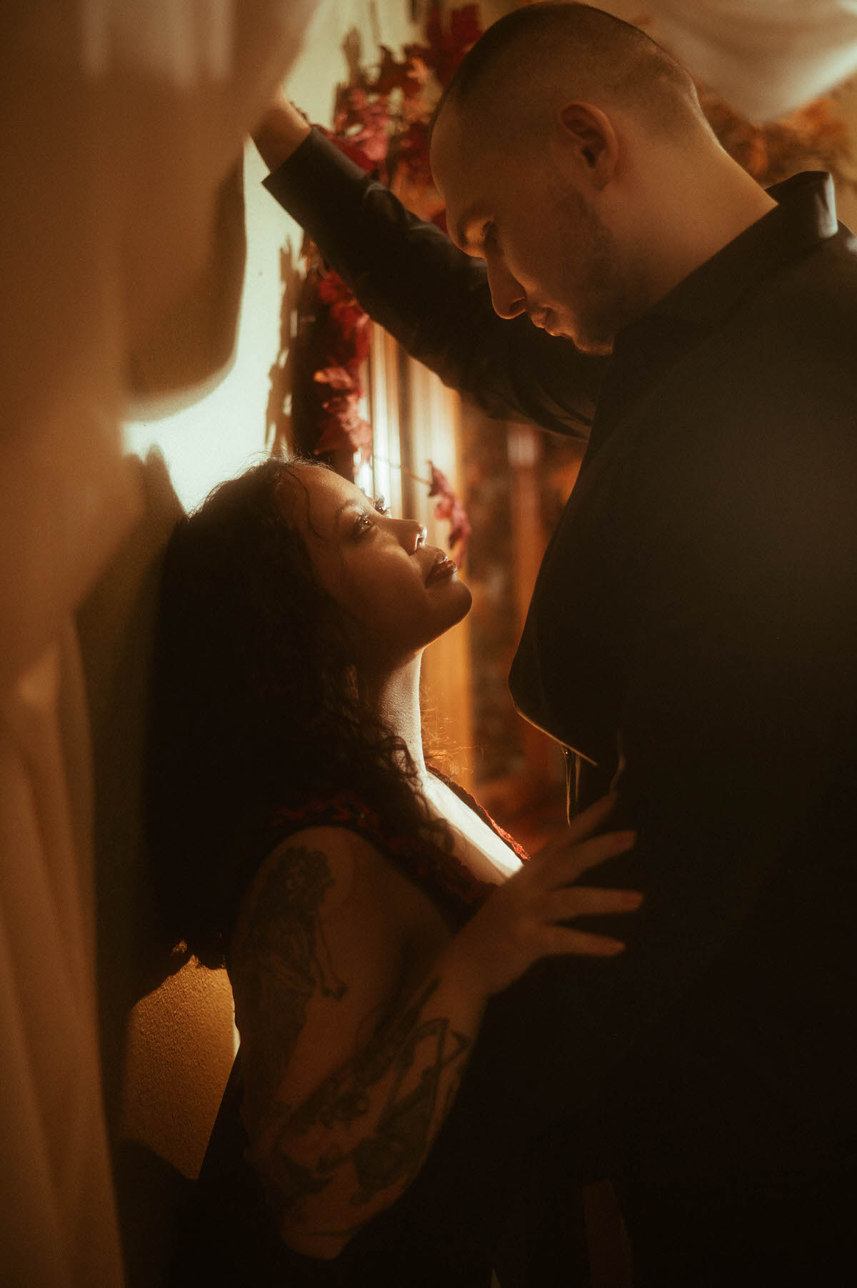 A woman leans against the wall gazing up at her partner, who towers over her with one arm pressed above her head. The golden candlelight casts soft shadows, adding drama and depth to this spicy couples boudoir moment, perfect for showcasing moody couples boudoir poses and romantic couples boudoir inspiration.