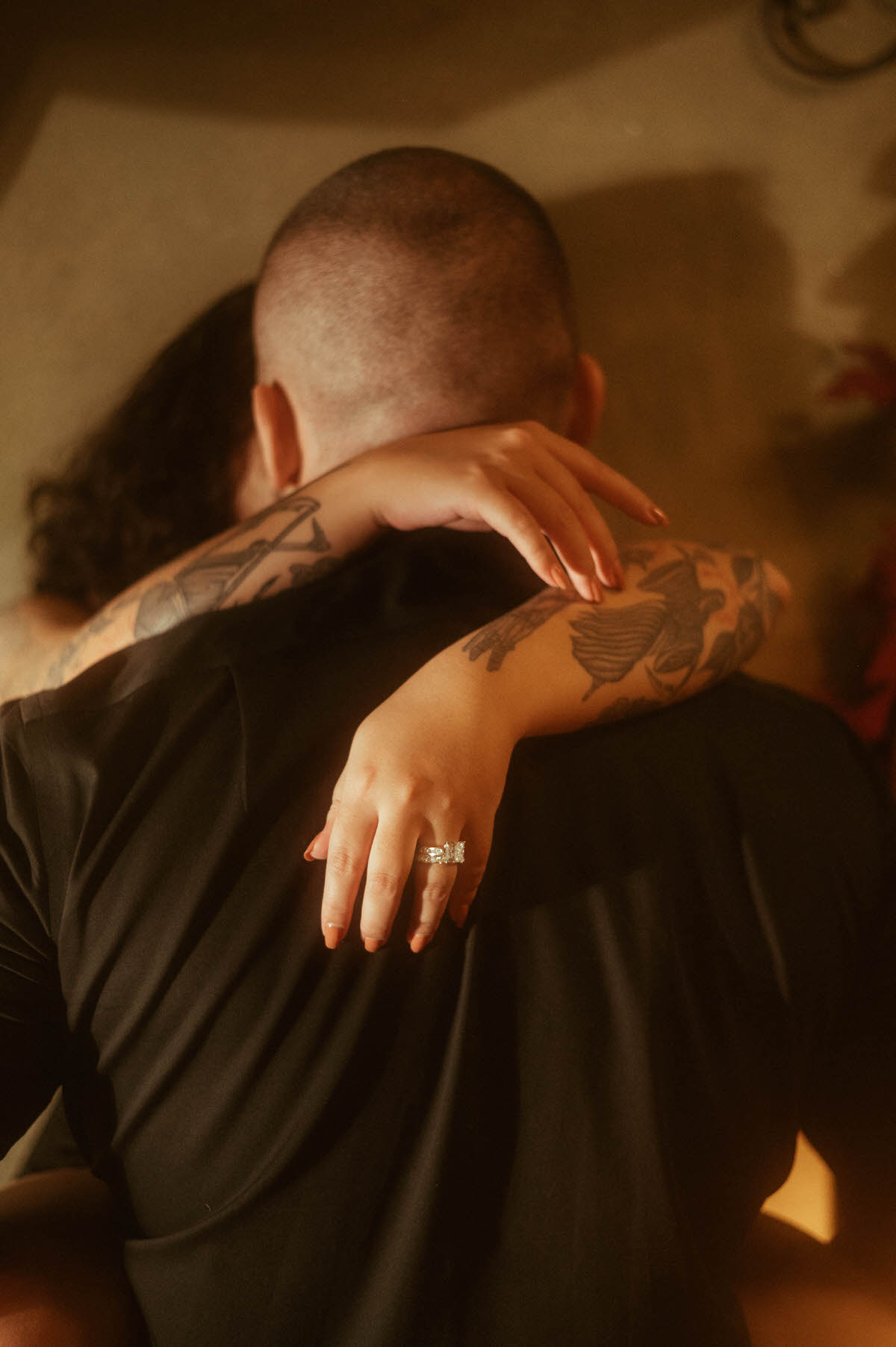 A close-up of a woman's tattooed arms wrapped around her partner's neck, her engagement ring glinting in the warm candlelight. This intimate embrace captures the essence of romantic couples boudoir and offers a subtle yet powerful take on moody couples boudoir ideas.