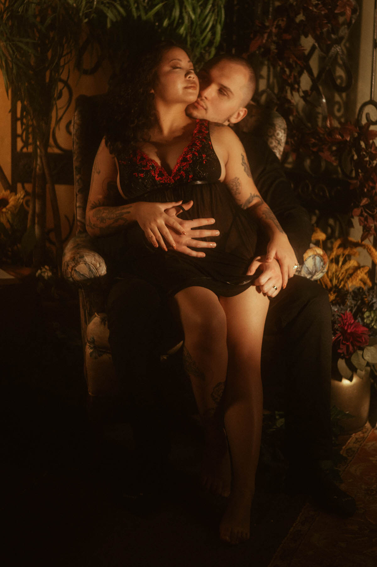 A woman sits in her partner’s lap on an ornate armchair, wearing a sheer black and red lingerie slip, as he wraps his arms around her waist and kisses her neck. This romantic couples boudoir portrait captures tender intimacy and sultry connection, offering moody couples boudoir inspiration and spicy couples boudoir pose ideas.