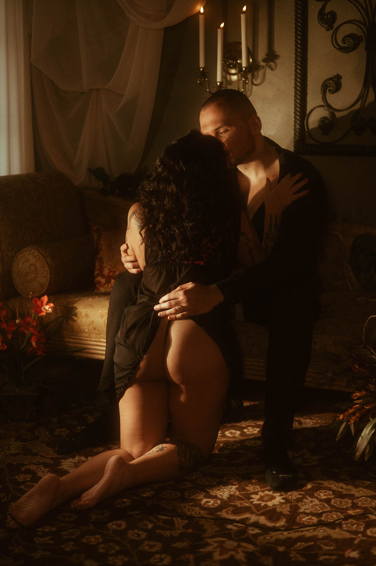 A woman kneels between her partner’s legs as they kiss in candlelight, framed by draped curtains and vintage decor. This romantic couples boudoir image evokes sensual connection and moody couples boudoir inspiration, perfect for those seeking bold and spicy couples boudoir ideas.
