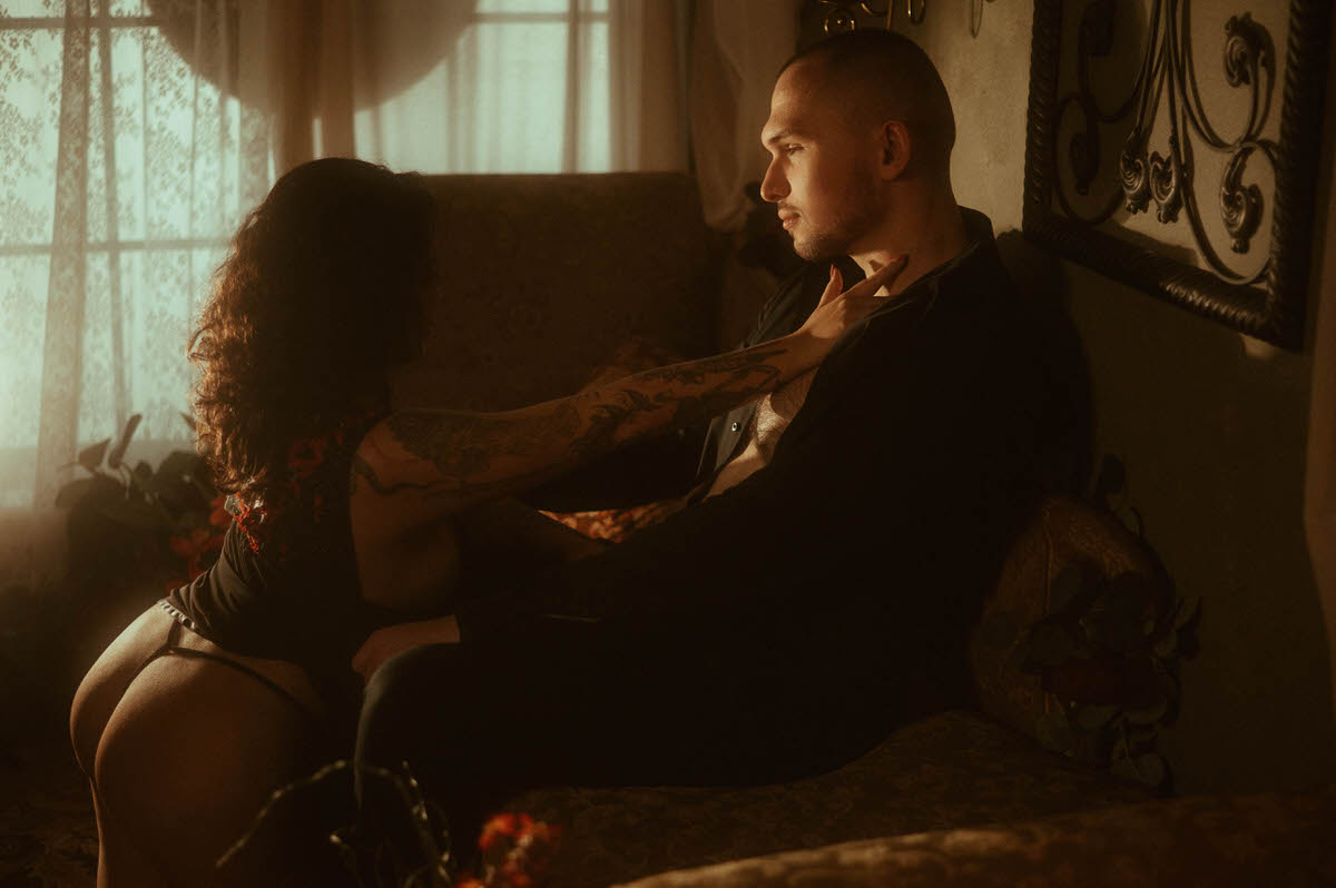A woman straddles her partner on a vintage sofa, reaching toward his chest as golden hour light filters through lace curtains. This romantic couples boudoir moment captures moody intimacy and connection, offering sultry inspiration for spicy couples boudoir poses and photo ideas.