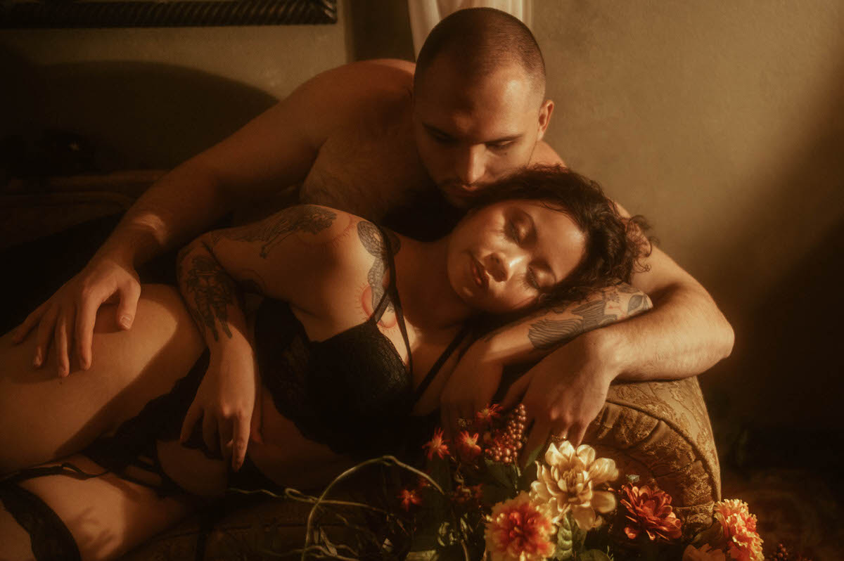 A couple lounges together on a brocade couch, the woman asleep in her partner’s embrace, wearing black lingerie. Warm lighting and a foreground of autumn florals create a soft, moody couples boudoir moment at Royal Lune Studio, ideal for romantic couples boudoir photography in Dallas.