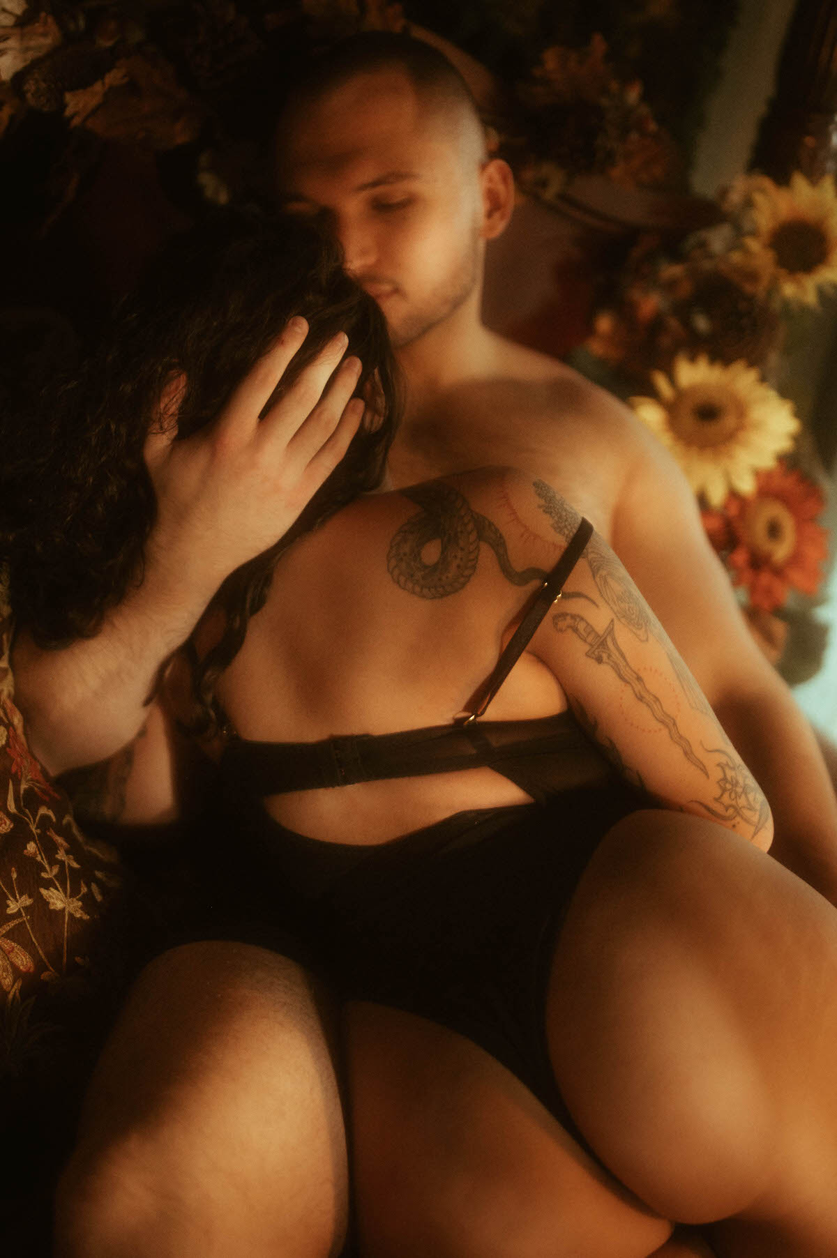 Sensual moment during a romantic couples boudoir session at Royal Lune Studio, featuring a woman in black lingerie straddling her partner as he gently cradles her head, surrounded by warm-toned florals and vintage decor.