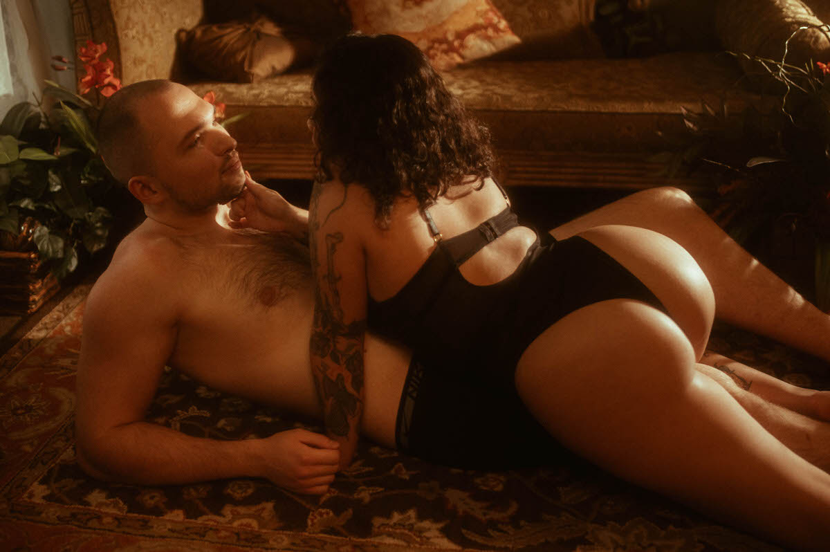 A romantic couples boudoir moment captured at Royal Lune Studio, where the woman straddles her partner on a vintage rug in soft golden light. Surrounded by antique furniture and dreamy shadows, the pose conveys intimacy and connection, perfect for a moody couples boudoir or spicy couple photoshoot.