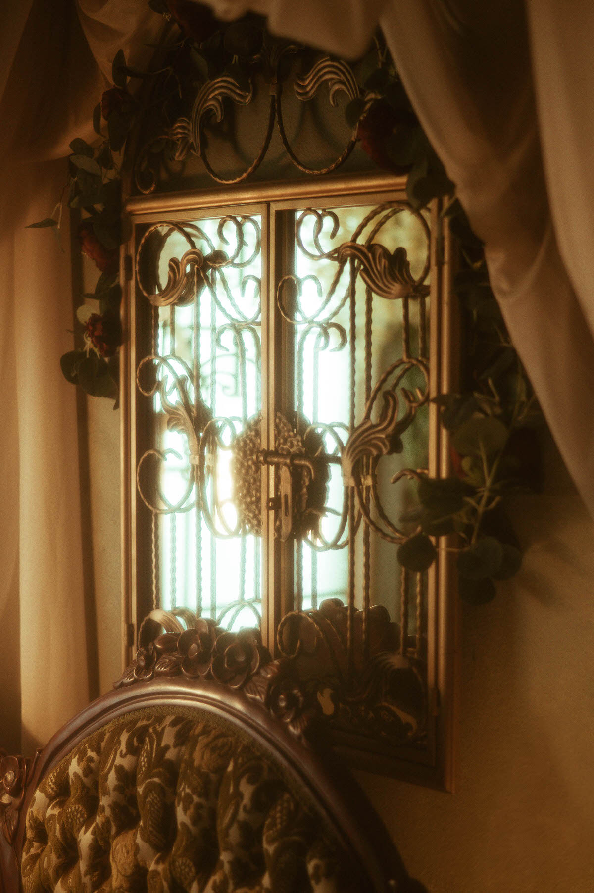 Close-up of an ornate gold metal window panel with scrollwork detail, framed by soft beige drapery and faux eucalyptus garland, glowing with backlight in the vintage-inspired spring setup at Royal Lune Studio, a Dallas boudoir studio.
