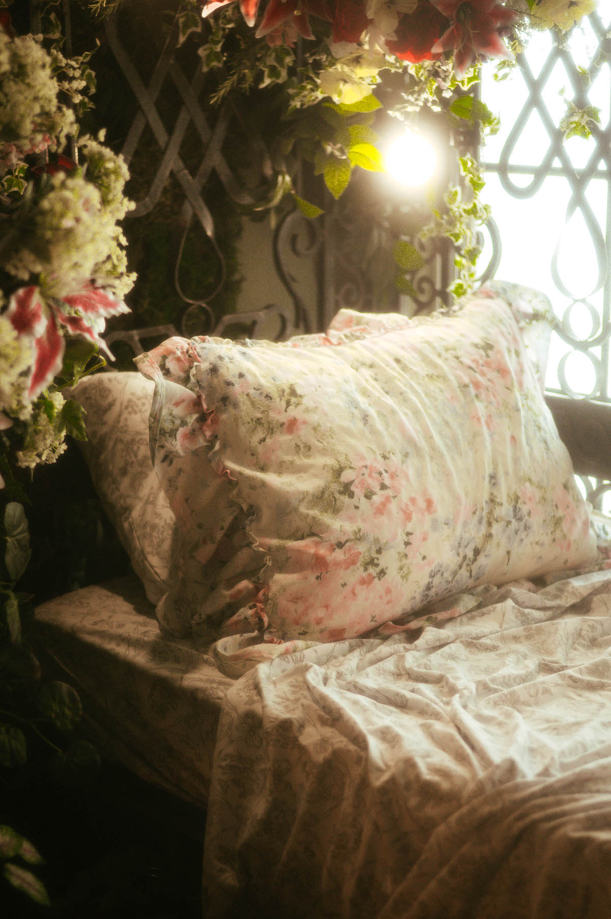Close-up of a romantic floral pillow and soft printed sheets on a daybed at Royal Lune Studio, surrounded by vines, soft light, and vintage wrought iron—capturing an ethereal spring boudoir studio setup in Dallas.