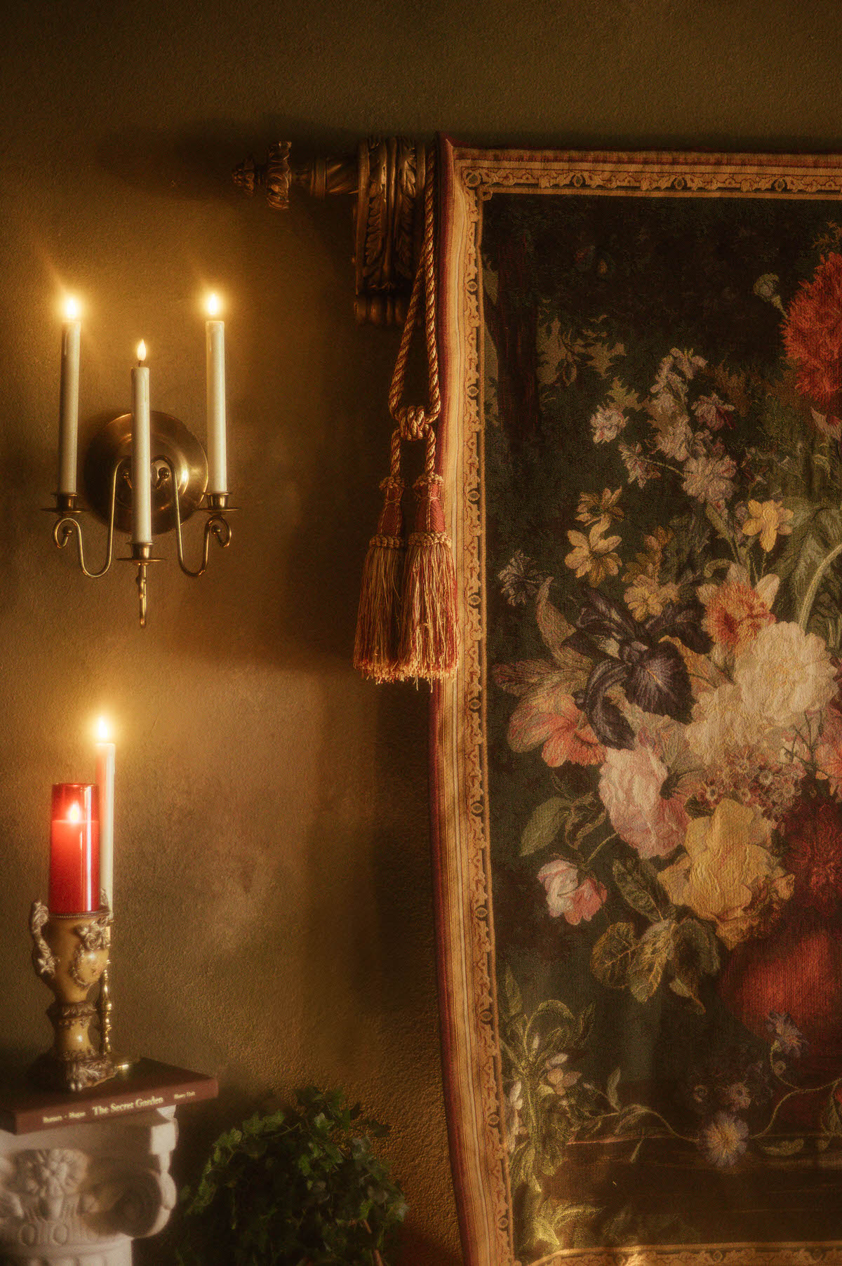 Close-up of a vintage floral tapestry with gold fringe tassel at Royal Lune Studio, accented by candlelight, ivy, and a bronze sconce on textured olive faux limewash walls in this Dallas boudoir studio.