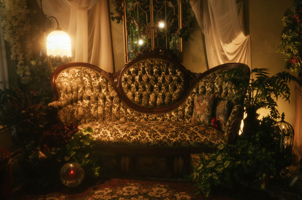 Victorian-style velvet sofa at Royal Lune Studio surrounded by lush greenery, vintage pillows, and soft golden lighting, with a beaded floor lamp and romantic drapery creating a dreamy spring boudoir studio setup in Dallas.