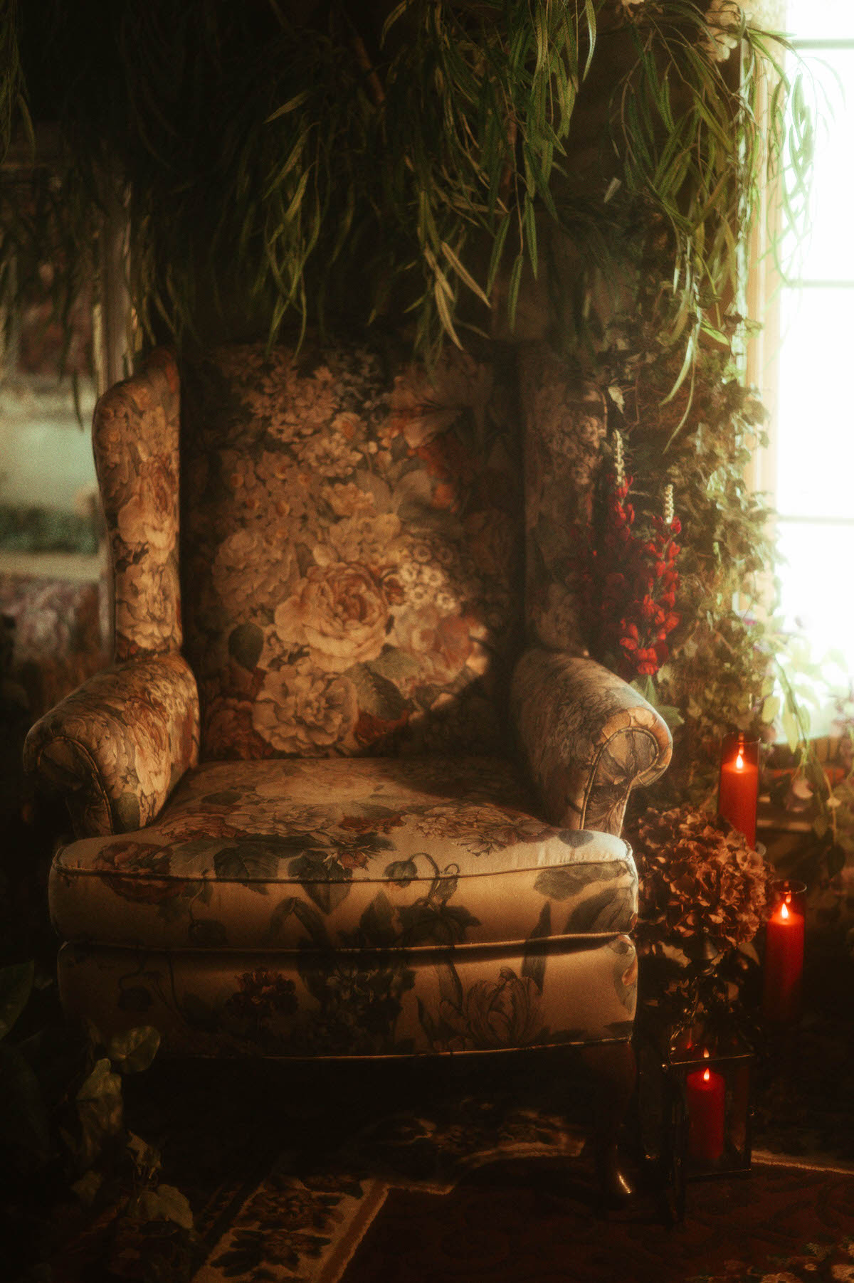 A vintage floral armchair bathed in soft golden light, surrounded by hanging greenery, red candles, and dried florals at Royal Lune Studio, part of a spring-inspired boudoir studio setup in Dallas.
