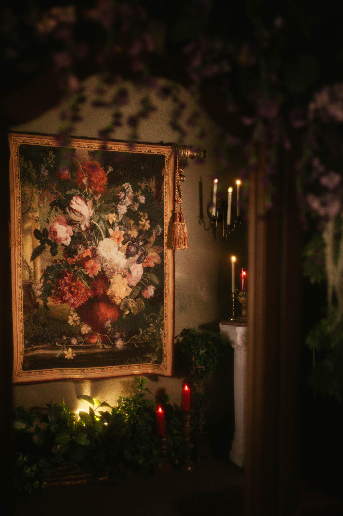 Romantic floral tapestry hangs on a faux limewash wall at Royal Lune Studio, surrounded by greenery, gold sconces, red candles, and classical column decor in this Dallas boudoir studio.
