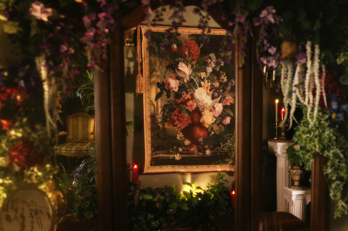 Reflection of a vintage floral still life tapestry framed by hanging greenery and faux wisteria at Royal Lune Studio, with red candles and classical columns adding to the romantic spring boudoir studio setup.