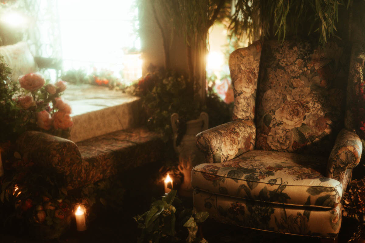 Sunlight floods through the window of Royal Lune Studio, illuminating a floral wingback chair and vintage daybed surrounded by pastel roses, greenery, and glowing candles in a spring boudoir studio setup in Dallas.
