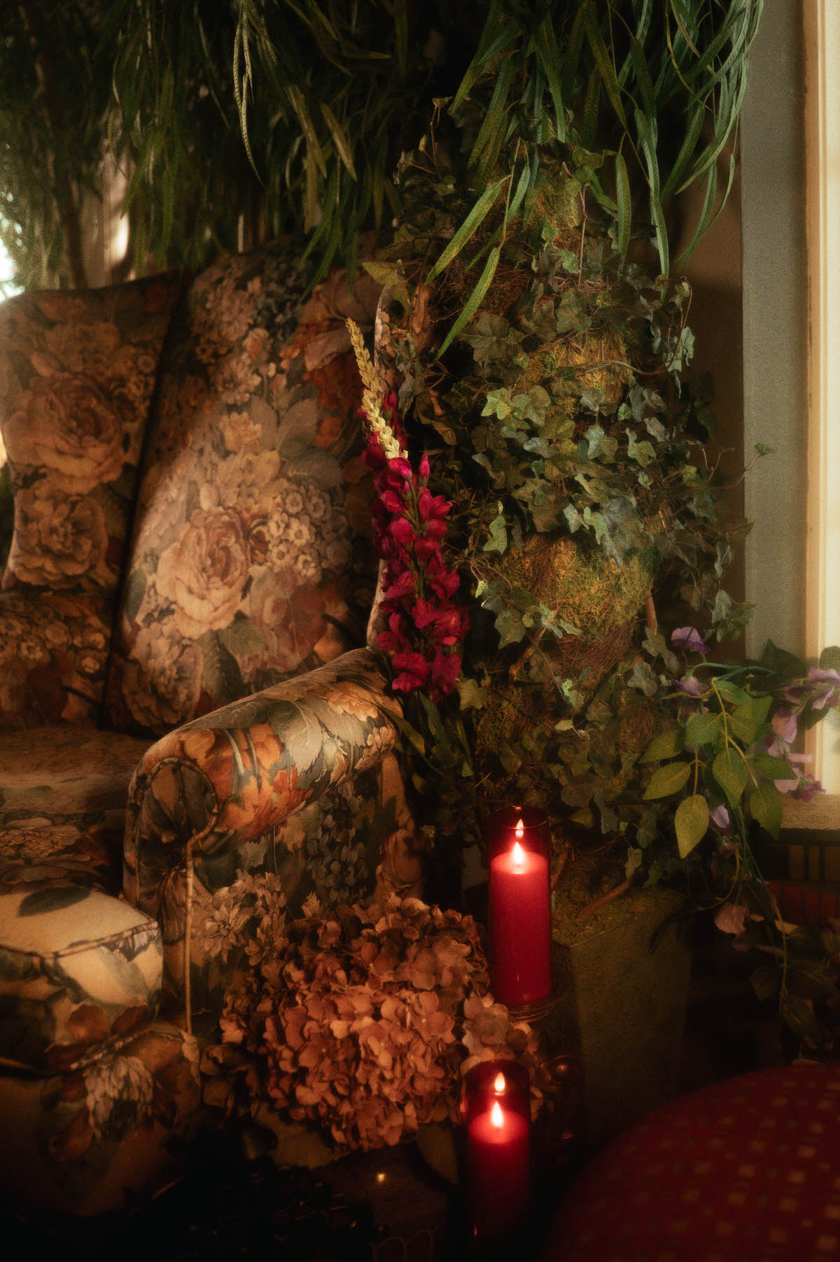 A floral tapestry armchair surrounded by cascading ivy, moss-covered decor, red pillar candles, and dried hydrangeas at Royal Lune Studio, styled for a romantic spring boudoir session in a Dallas photography studio.