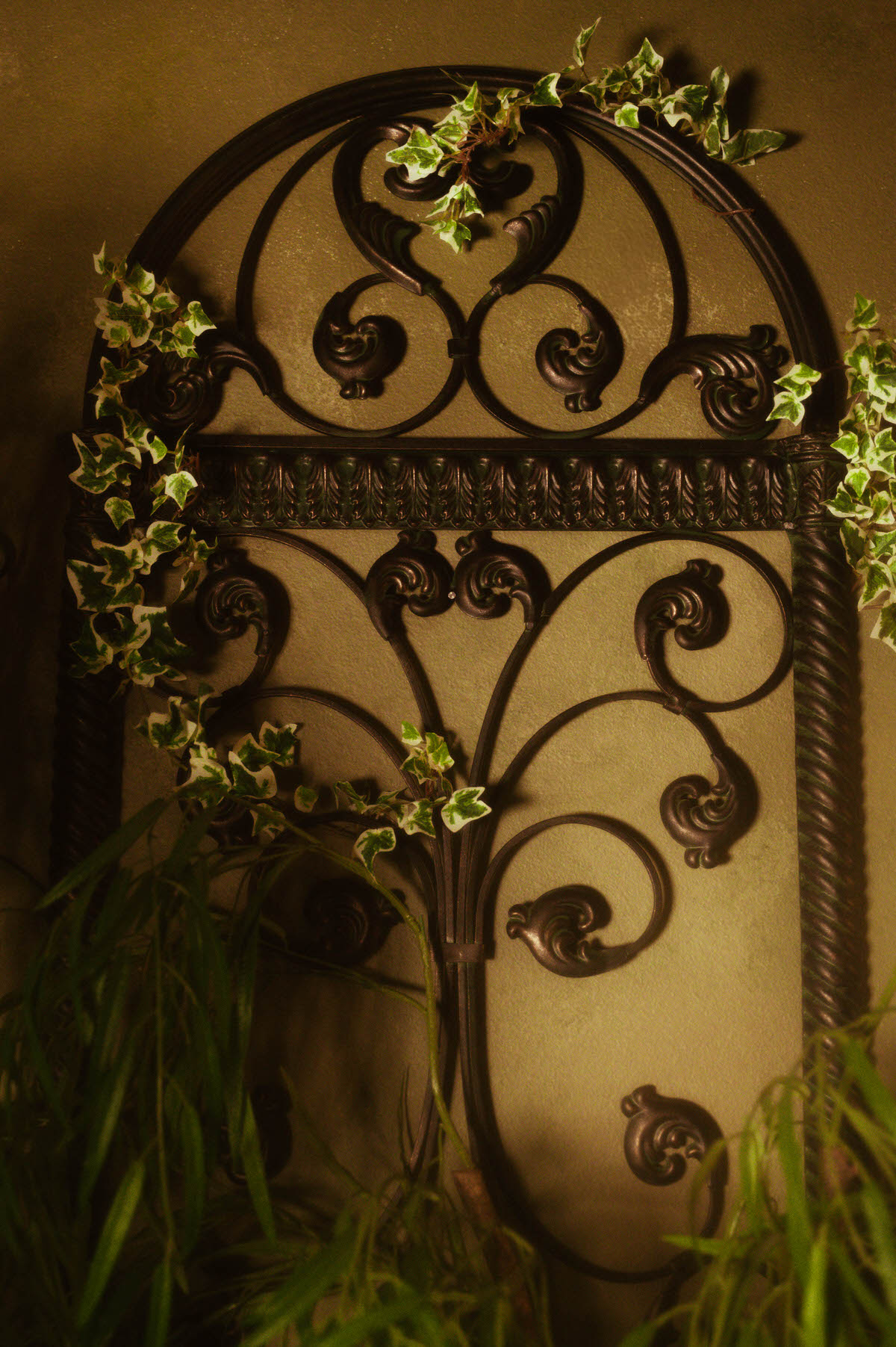 Close-up of a vintage wrought iron headboard with ornate scrollwork, styled with trailing faux ivy against the olive-toned limewash wall at Royal Lune Studio, a dreamy Dallas boudoir studio.