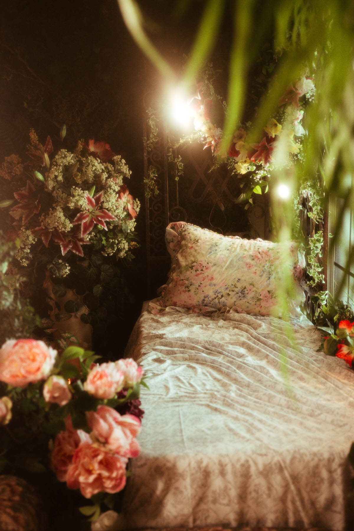 Floral daybed styled with a pastel pillow and botanical bedsheets, lit by warm bulbs and framed with ivy, lilies, and trailing blooms—part of the enchanted spring setup at Royal Lune Studio, a Dallas boudoir studio.