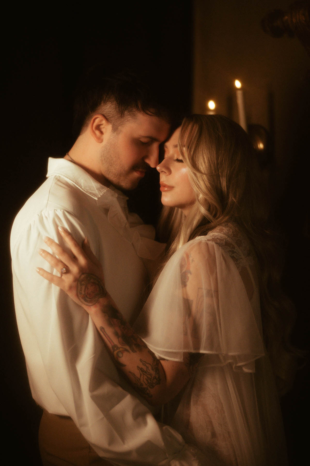 A tender engagement photo of a couple with their foreheads touching, lit by candlelight at Royal Lune Studio. The bride-to-be wears a sheer vintage-style lace robe, showcasing her engagement ring, while the groom-to-be wears a white ruffled shirt. This intimate moment evokes dreamy engagement photography with romantic couples photography styling and artistic couple posing, perfect for Dallas wedding photography inspiration.