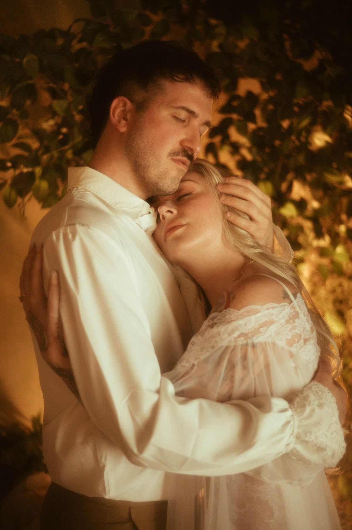 A romantic moment from a Dallas engagement photos session, where the couple embraces with eyes closed in soft candlelight. The woman rests her head tenderly against her partner’s chest, wearing a sheer lace gown that adds a vintage feel. His ruffled shirt and their intimate pose capture the essence of dreamy engagement photography and artistic couple poses.