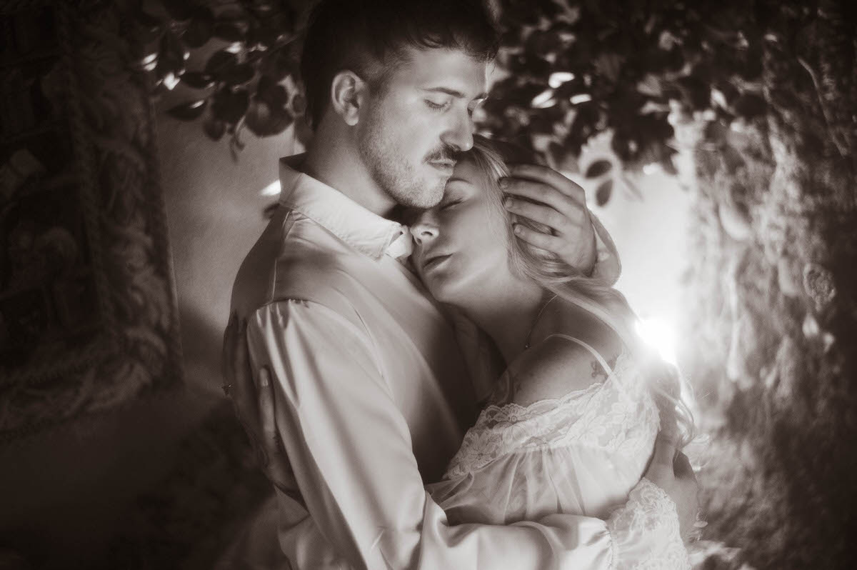 A black-and-white image from a Dallas engagement photos session, showing the couple wrapped in an emotional embrace. The woman rests her head on her partner’s chest with eyes closed, while he gently holds her head, evoking a tender and protective mood. Her vintage lace gown and his romantic shirt enhance the dreamy engagement photography style.