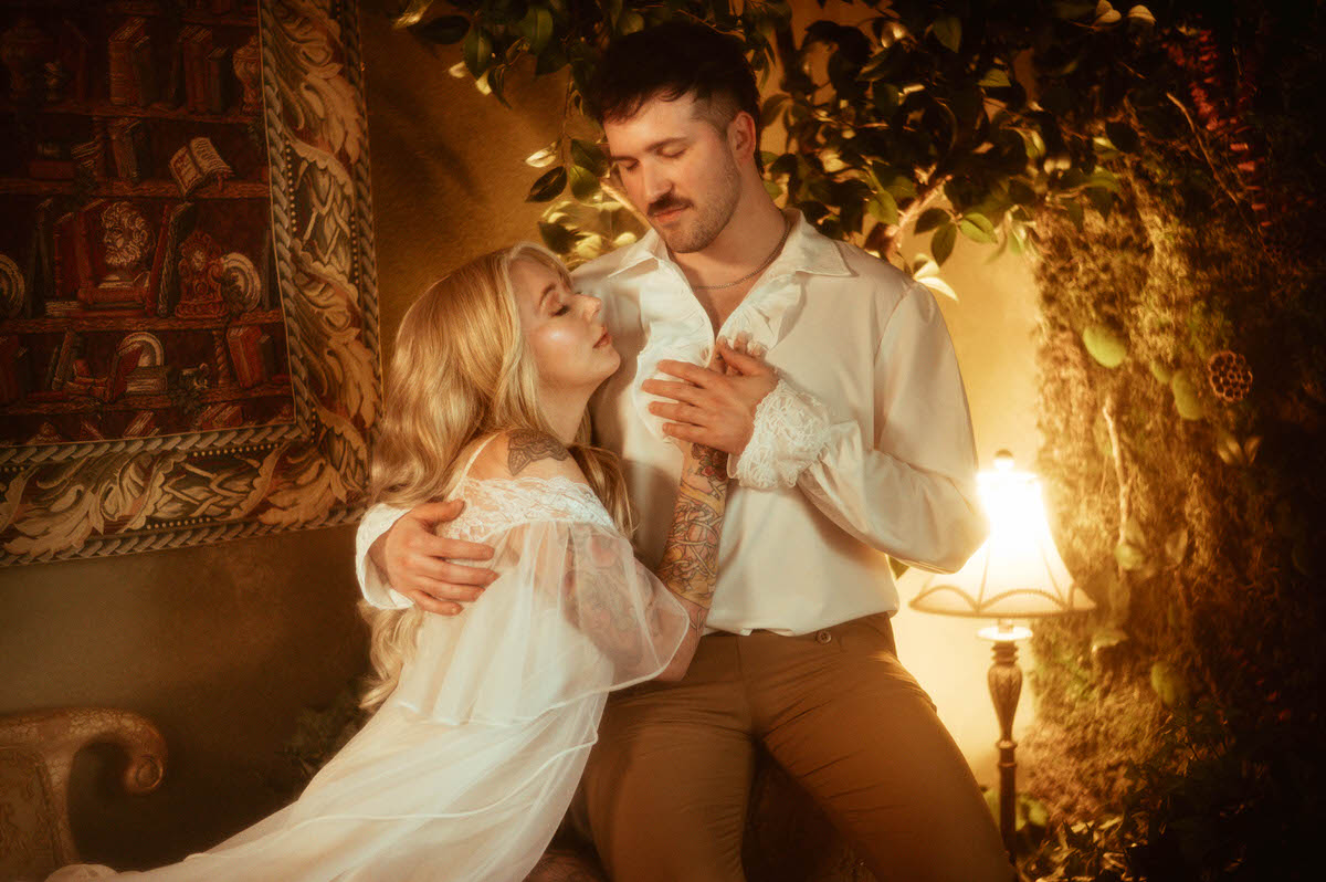 A dreamy engagement photo of a couple in an intimate pose at Royal Lune Studio, with the woman resting her head on her partner’s chest while holding his hand. The setting features vintage decor, including a tapestry, velvet seating, and a moss wall illuminated by soft golden light. Their Renaissance-inspired wardrobe and the romantic atmosphere highlight artistic couple poses and dreamy engagement photography for Dallas wedding photography inspiration.