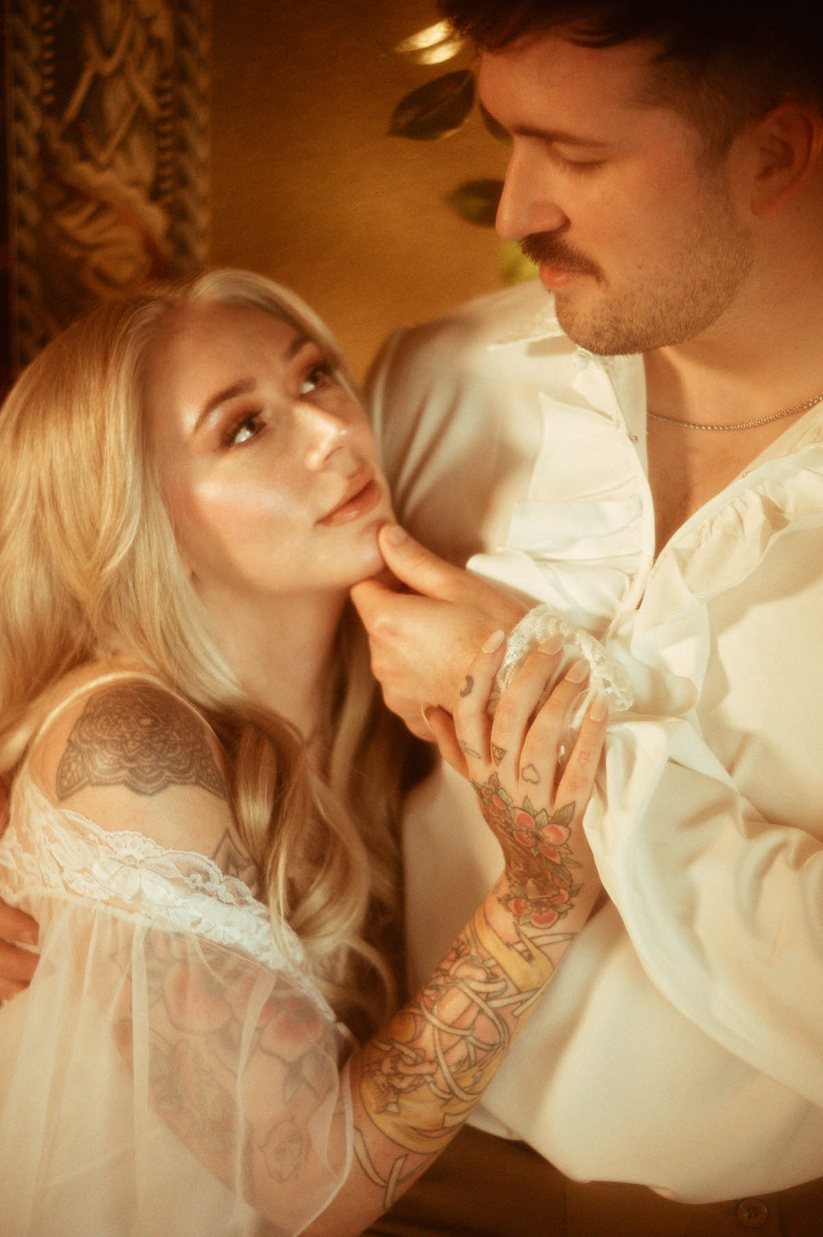 Close-up of a romantic couple during their Dallas engagement photos at Royal Lune Studio. The woman gazes lovingly at her partner while holding his hand, showing off her engagement ring and floral tattoos. Her sheer lace gown and his Renaissance-inspired shirt add to the dreamy engagement photography vibe, perfect for artistic couple poses and romantic wedding photography in Dallas.