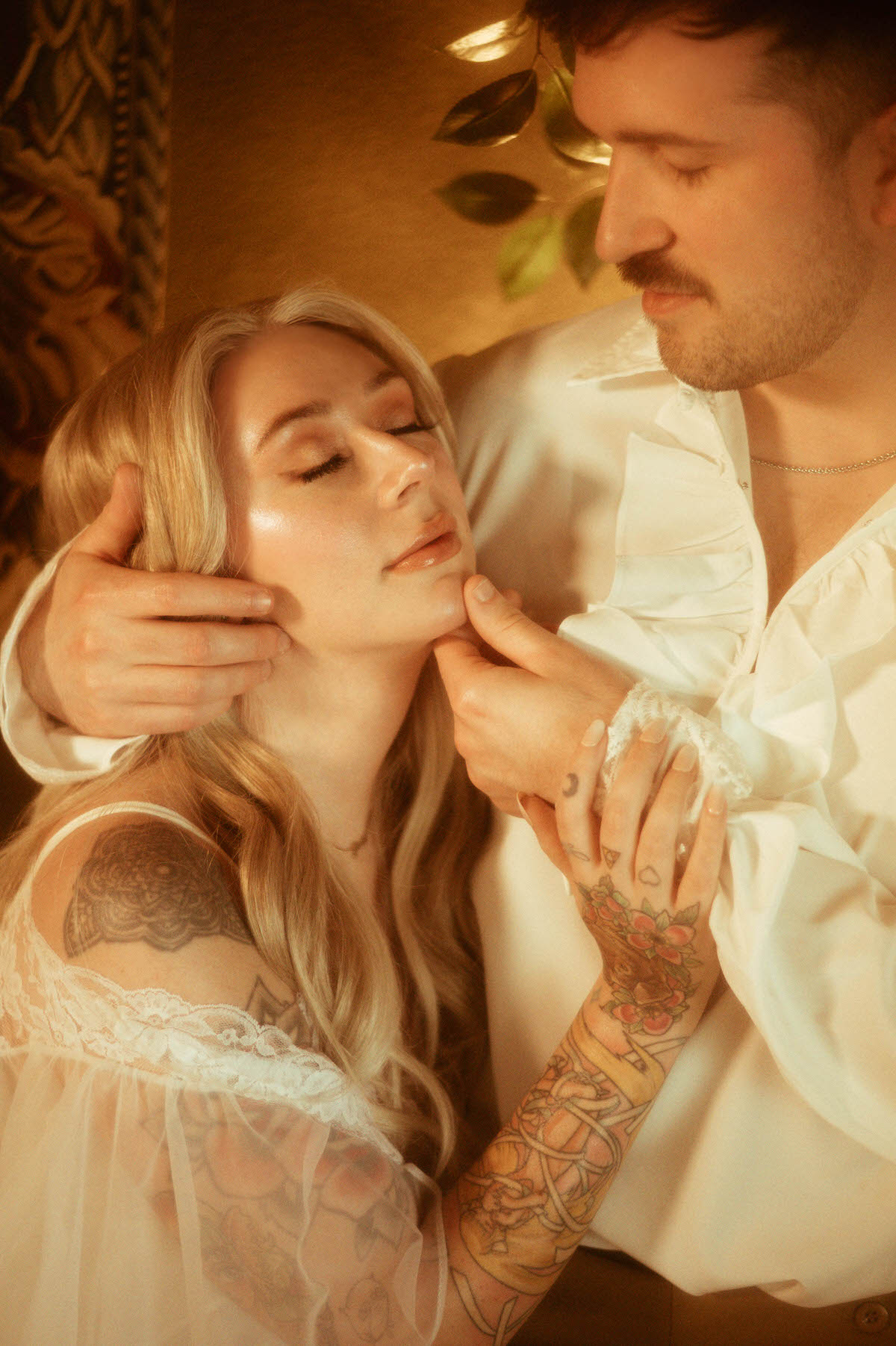 A tender close-up from a Dallas engagement photos session, capturing the woman with her eyes closed and face gently held by her partner. Her sheer lace gown, visible tattoos, and engagement ring highlight the romantic couples photography style, while his ruffled shirt nods to an artistic engagement photos outfit. The golden light adds to the dreamy engagement photography mood at Royal Lune Studio.