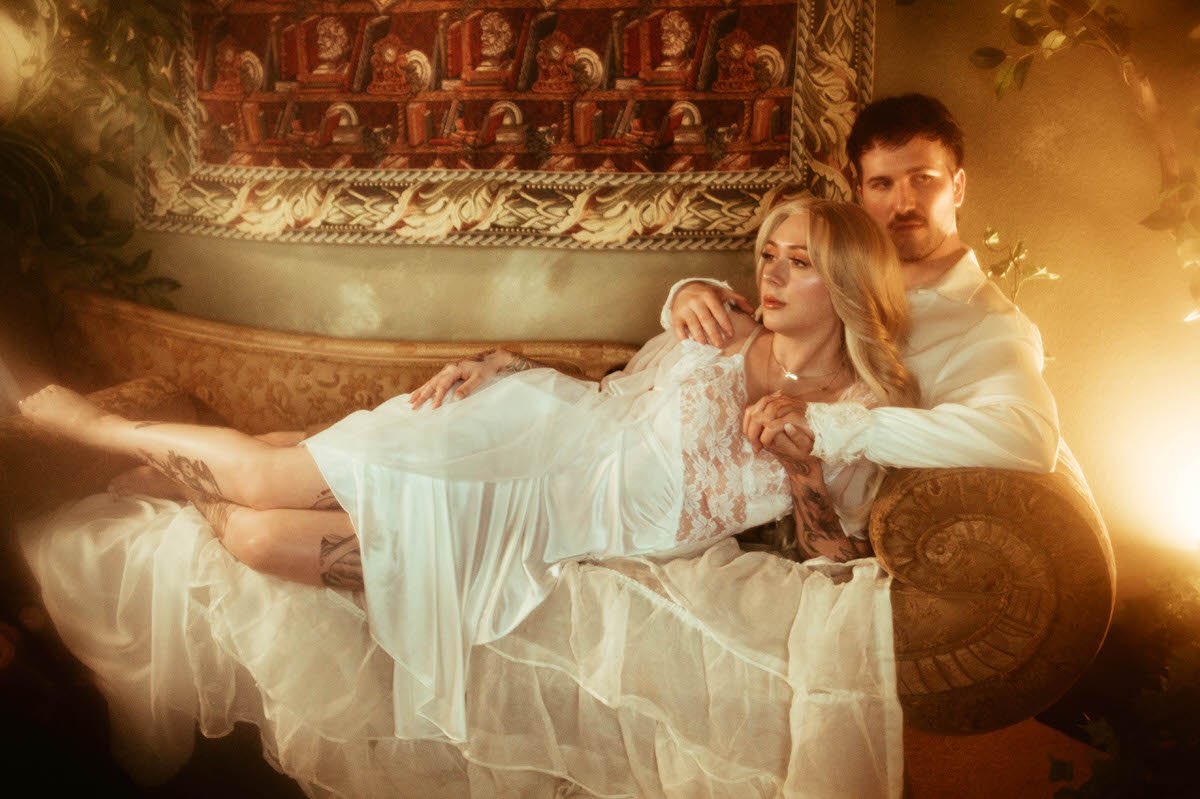 A couple poses for dreamy Dallas engagement photos, with the woman reclining across a vintage tapestry couch in a sheer lace nightgown while the man sits behind her, holding her hands. The romantic setting includes golden lighting, antique furniture, and lush greenery, capturing an artistic couple pose perfect for engagement photos outfit inspiration.