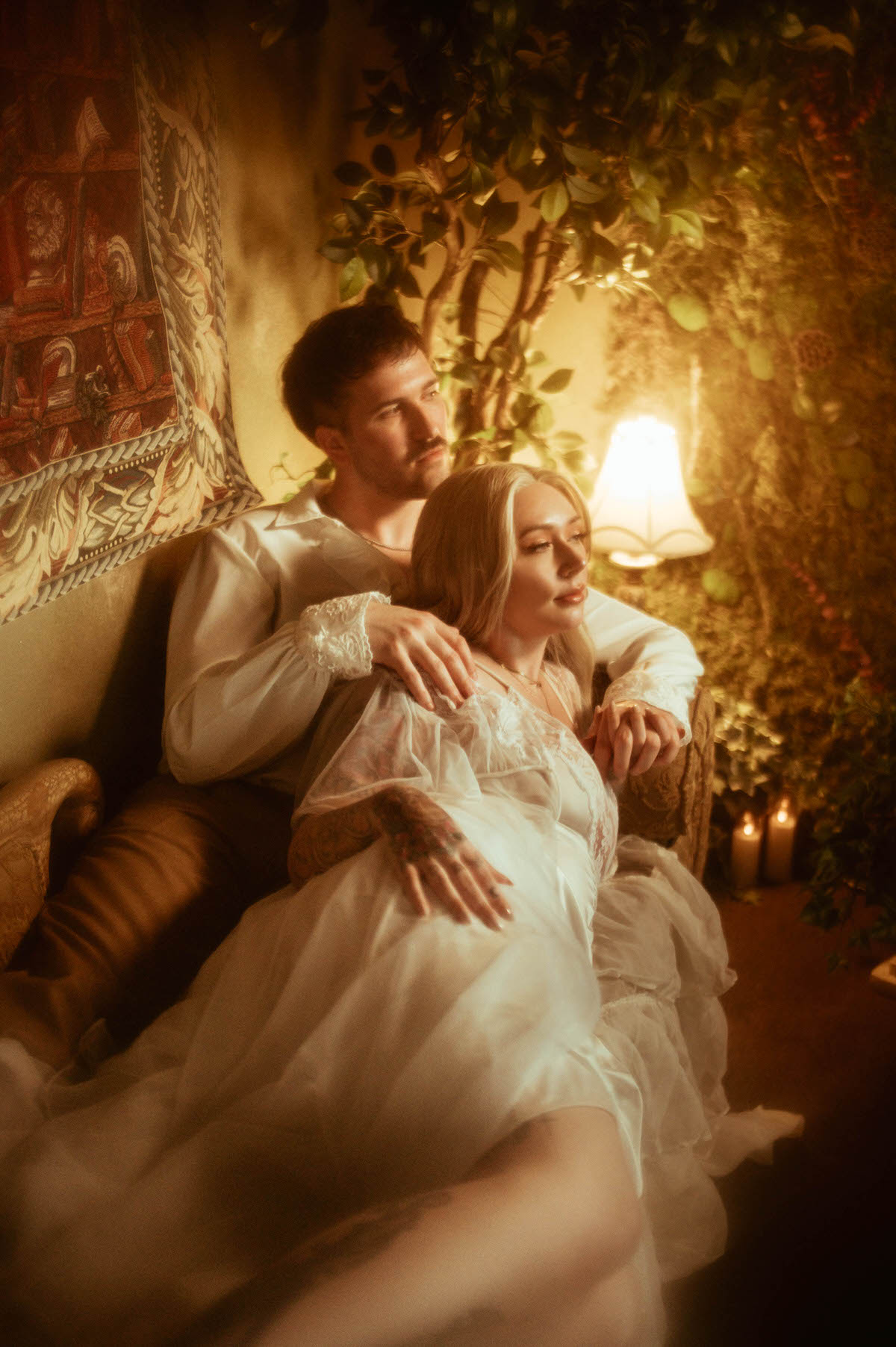 A romantic engagement photo featuring a couple lounging on a vintage tapestry couch, with the woman resting against the man in a sheer lace gown. Warm golden lighting, flickering candles, and lush greenery create a cozy, dreamy setting inside Royal Lune Studio, perfect for artistic Dallas engagement photos.