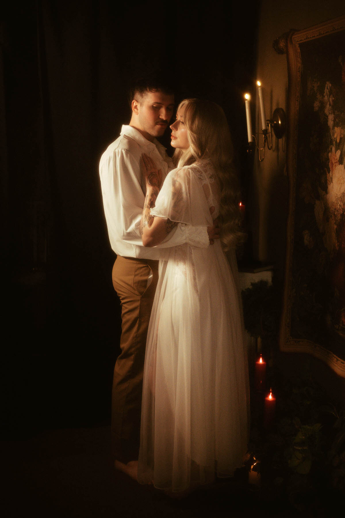 A romantic engagement portrait at Royal Lune Studio featuring a couple embracing beside candlelight and a floral tapestry. The bride-to-be wears a flowing sheer gown, while her partner wears a ruffled shirt and tan pants. This image captures the soft intimacy of dreamy engagement photography with vintage-inspired engagement photo outfits and artistic couple poses in a moody, candlelit setting.