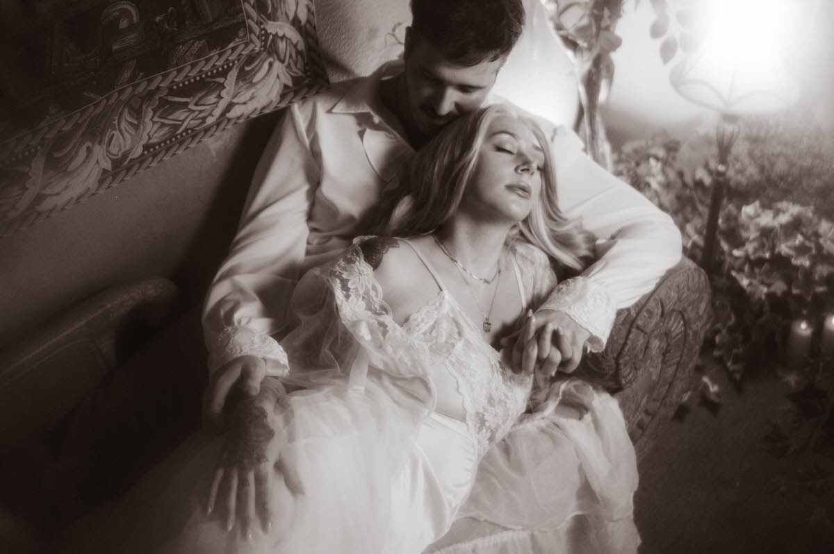 A black and white engagement photo of a couple embracing on a vintage couch at Royal Lune Studio. The woman, in a lace bridal-style gown, leans back against her partner with her eyes closed, creating a dreamy, intimate moment amid candlelight, ivy, and antique decor. Perfect for romantic Dallas engagement photography.