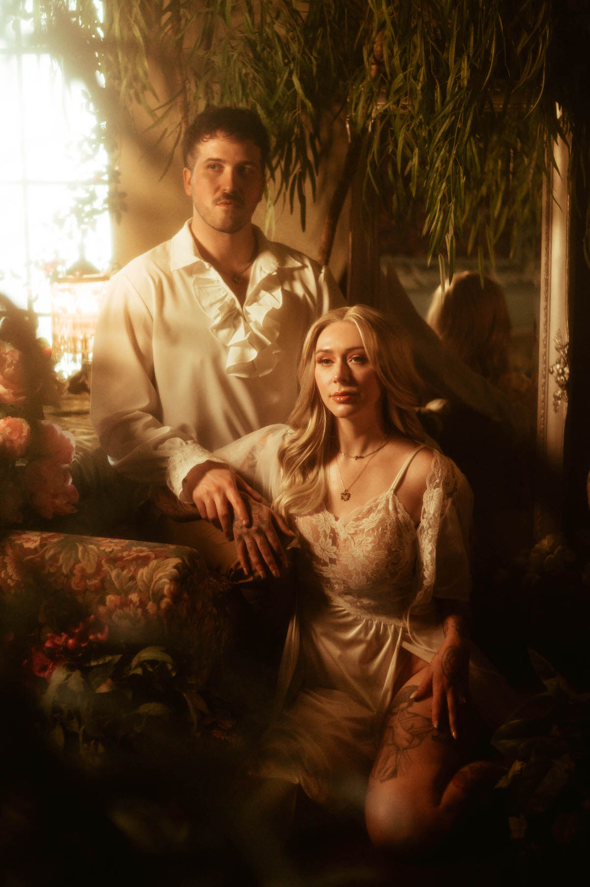 Romantic engagement portrait of a couple in vintage attire at Royal Lune Studio, with the woman kneeling in a lace nightgown beside her partner, who stands behind her with his hand resting on hers. Surrounded by soft floral textures, warm golden light, and a dreamy mirror reflection, this Dallas engagement photo blends fantasy and fine art elegance.
