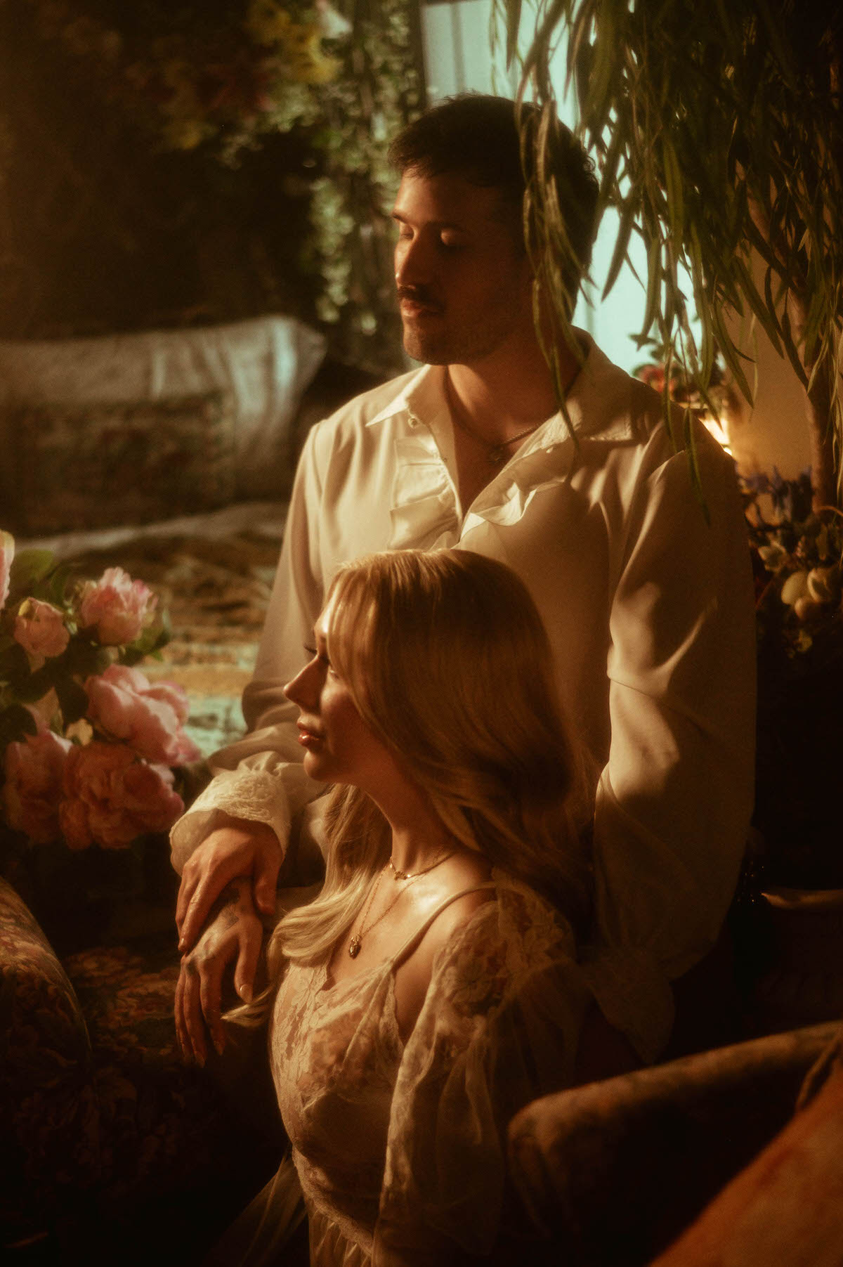 Romantic engagement portrait at Royal Lune Studio featuring a couple in vintage-inspired outfits, softly lit by golden window light. The woman sits in profile wearing a sheer lace gown, while her partner stands behind her, his hand resting tenderly on hers. The setting is adorned with lush florals and antique textures, evoking a dreamy and ethereal mood for Dallas engagement photos.