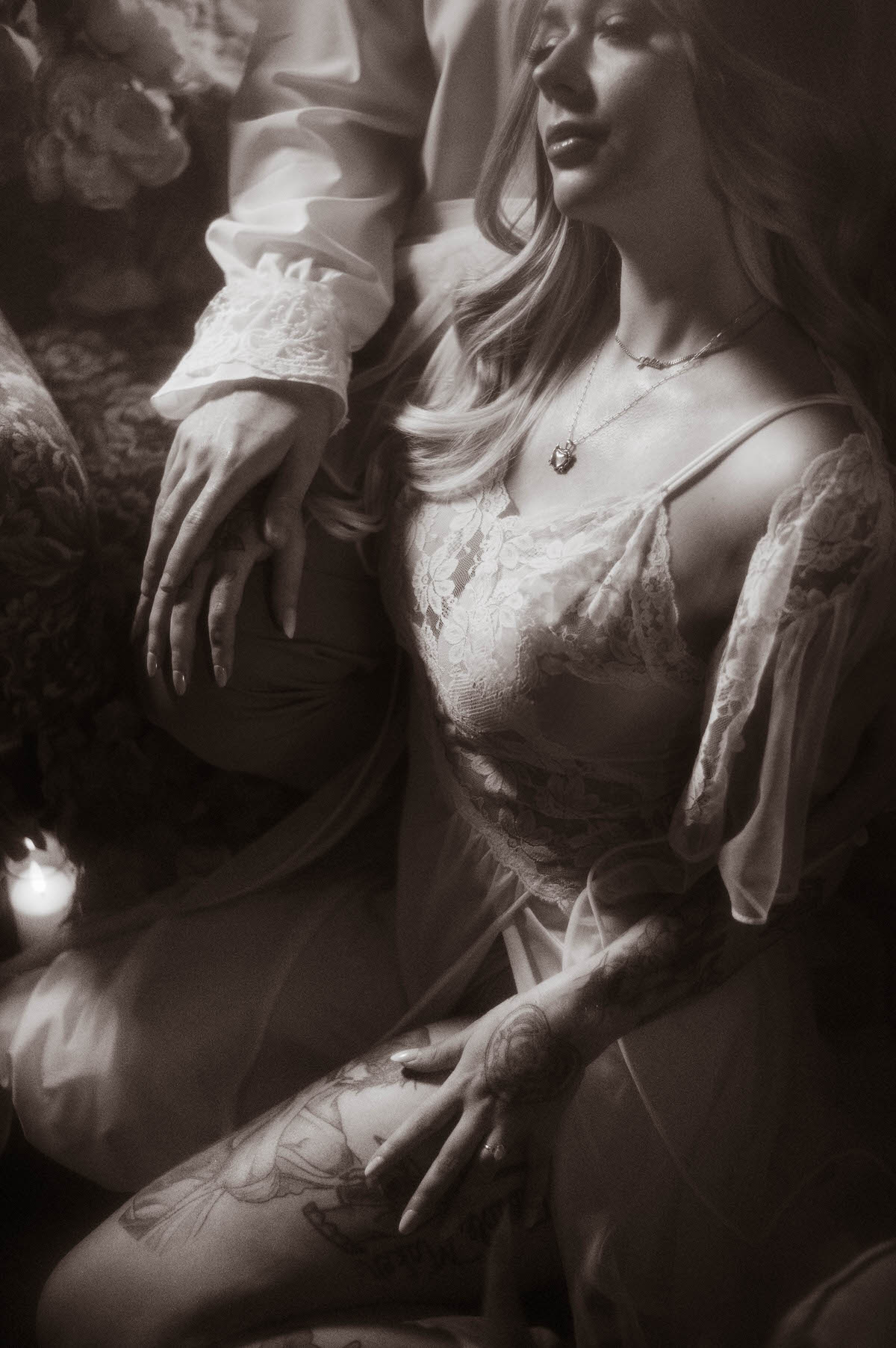 Black and white close-up from a Dallas engagement session at Royal Lune Studio, capturing the bride-to-be in a sheer lace gown as she rests against her partner’s leg. Her hand, adorned with an engagement ring, gently touches her thigh, highlighting detailed tattoos and soft candlelight for a vintage, romantic atmosphere.