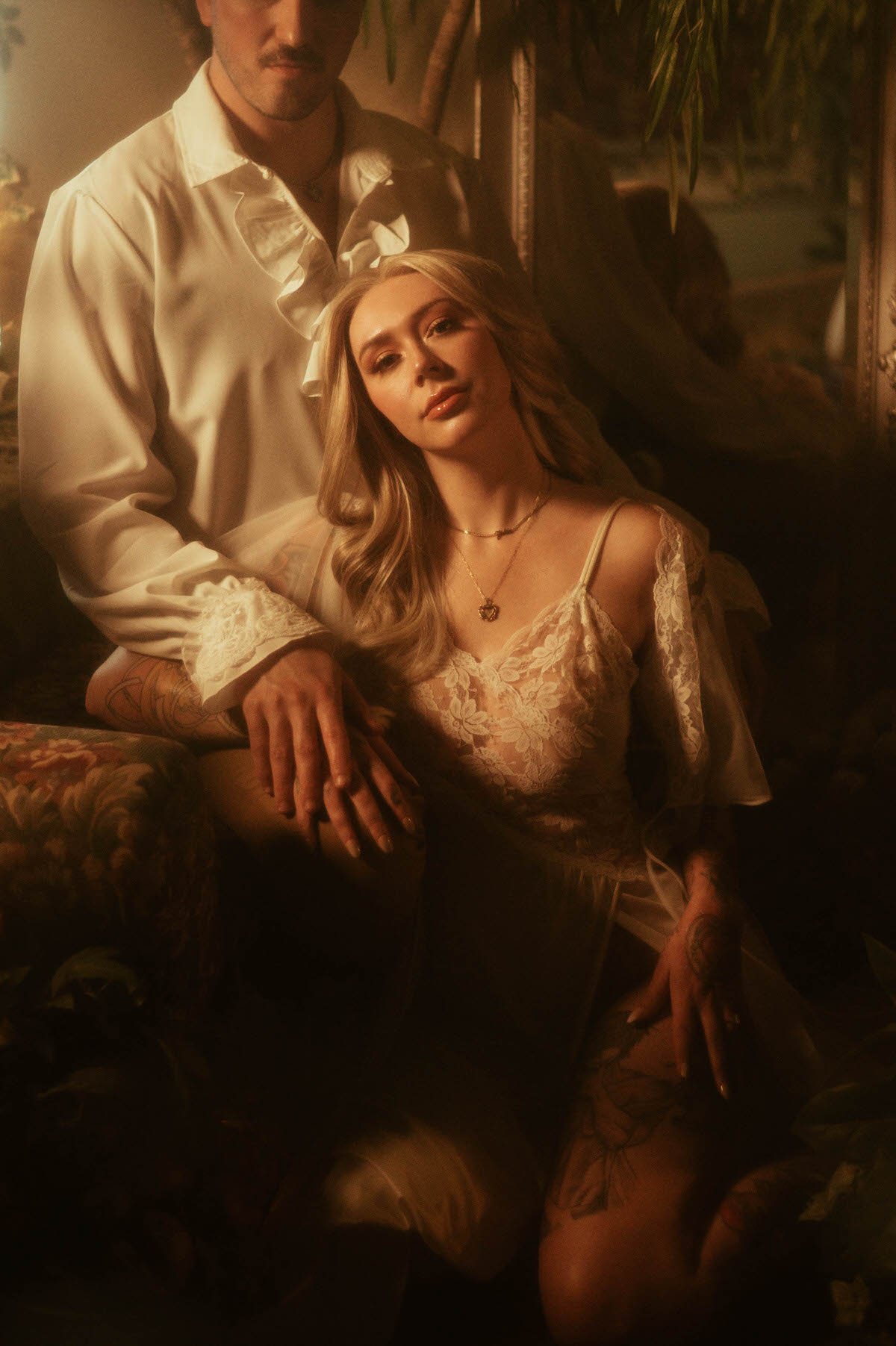 Intimate engagement portrait at Royal Lune Studio featuring a couple in vintage-inspired attire, with the woman kneeling gracefully in a sheer lace gown while leaning against her partner's leg. Surrounded by romantic florals and soft golden light, this Dallas engagement photo evokes a dreamy, painterly atmosphere.